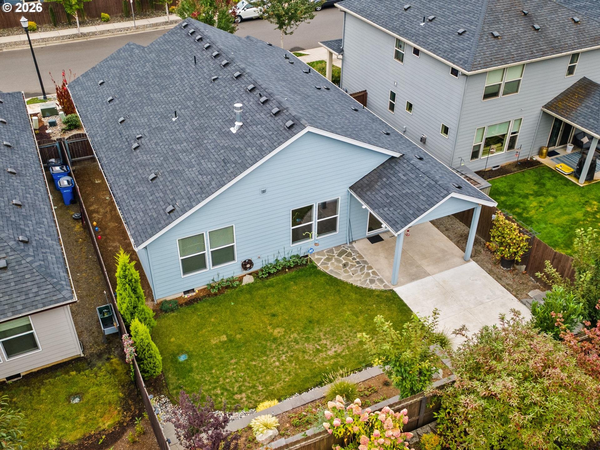 Aerial view of a light-blue home’s backyard, covered patio, lawn, and flowering garden.
