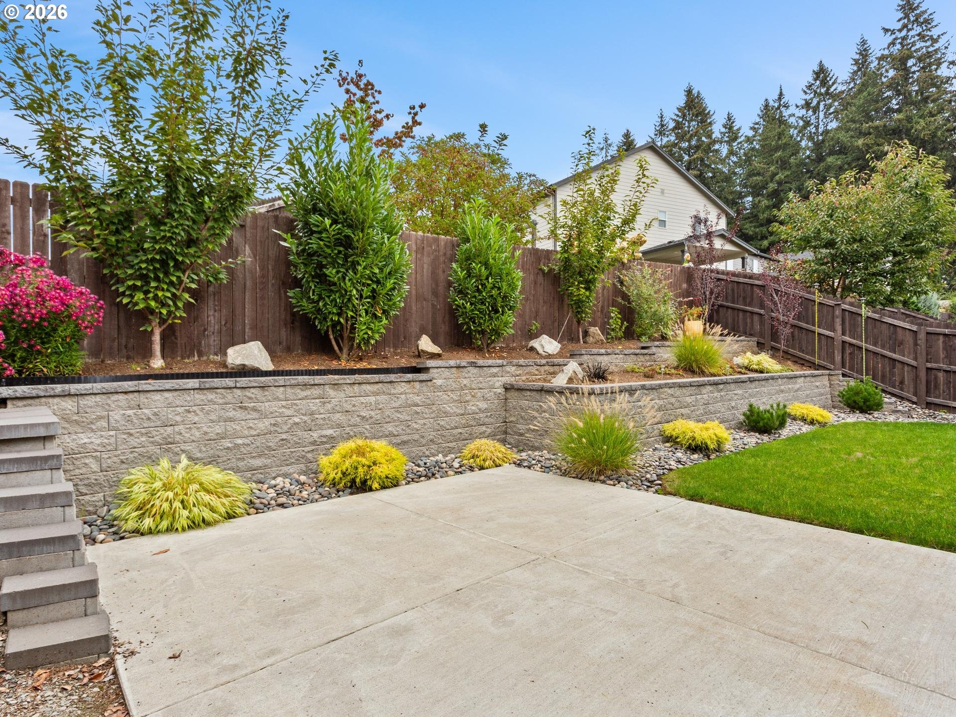 Backyard patio with tiered stone retaining walls, colorful landscaping, lawn, and a wooden privacy fence.
