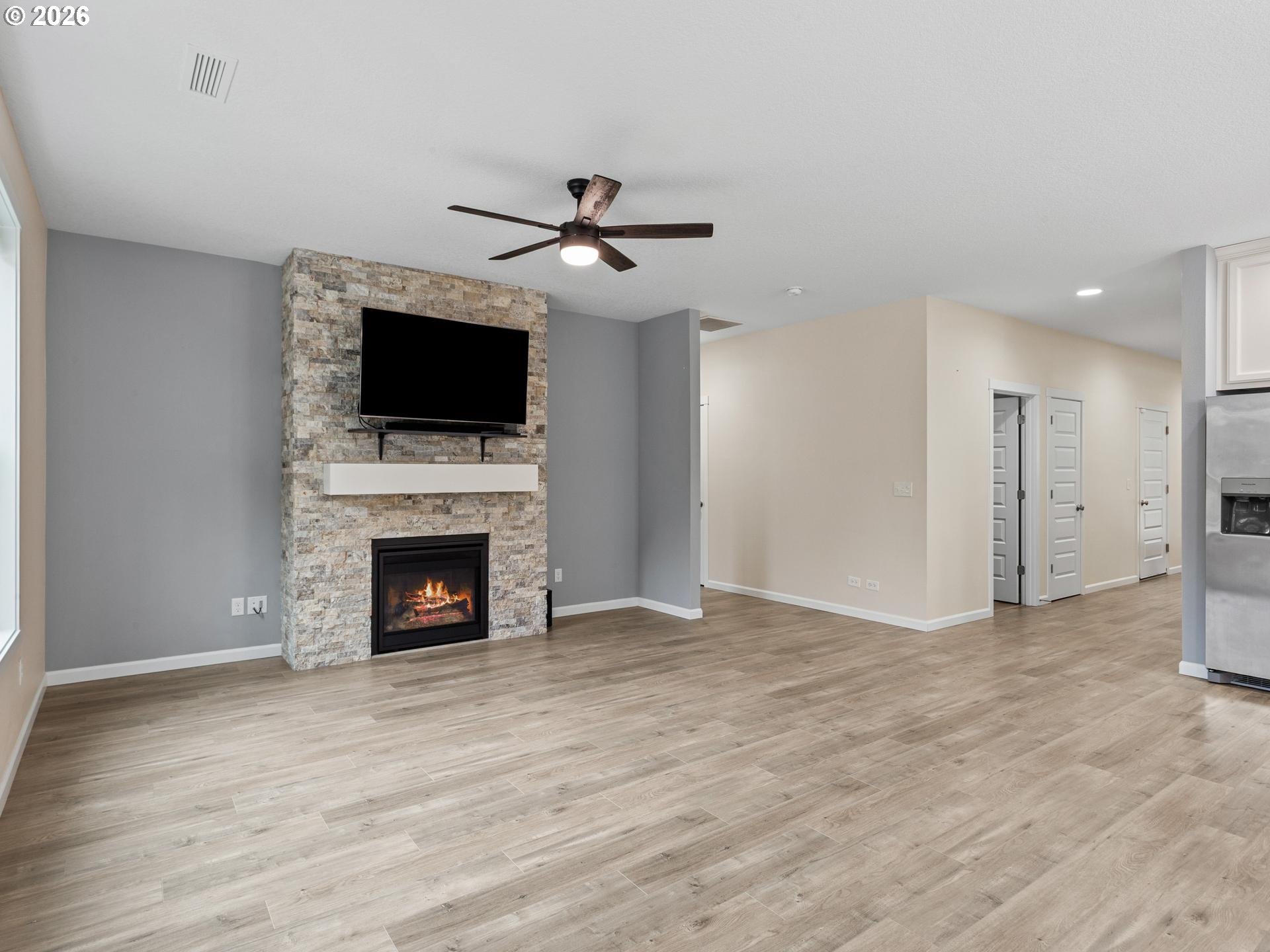 Unfurnished living room with a stone fireplace, mounted television, ceiling fan, and light wood-look flooring.