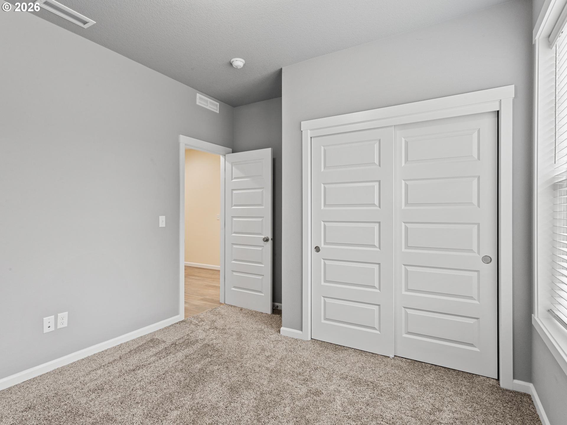 Empty bedroom with beige carpet, white sliding closet doors, and a window with blinds.