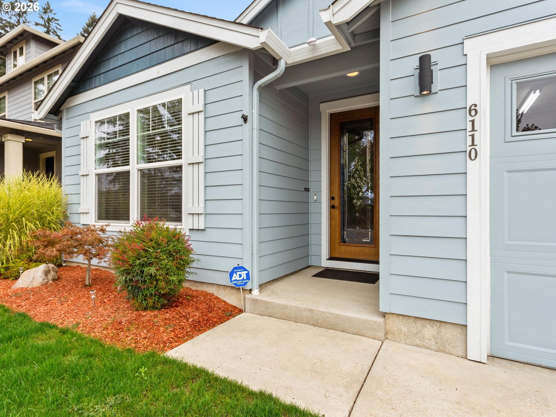 Light blue house entrance with a covered porch, wood-framed glass door, and landscaped front garden.
