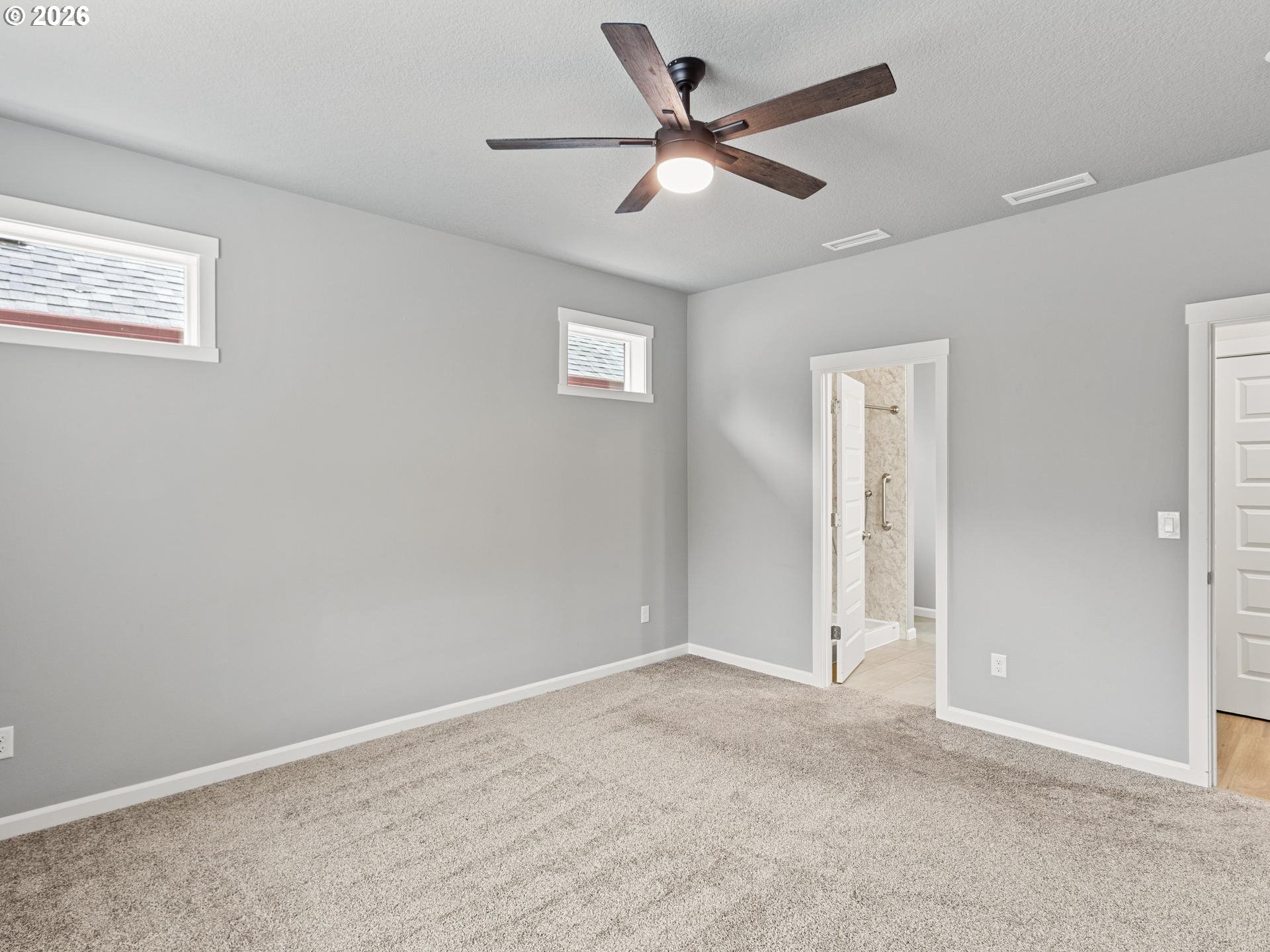 Empty carpeted bedroom with gray walls, high windows, and a ceiling fan; bathroom doorway is open.