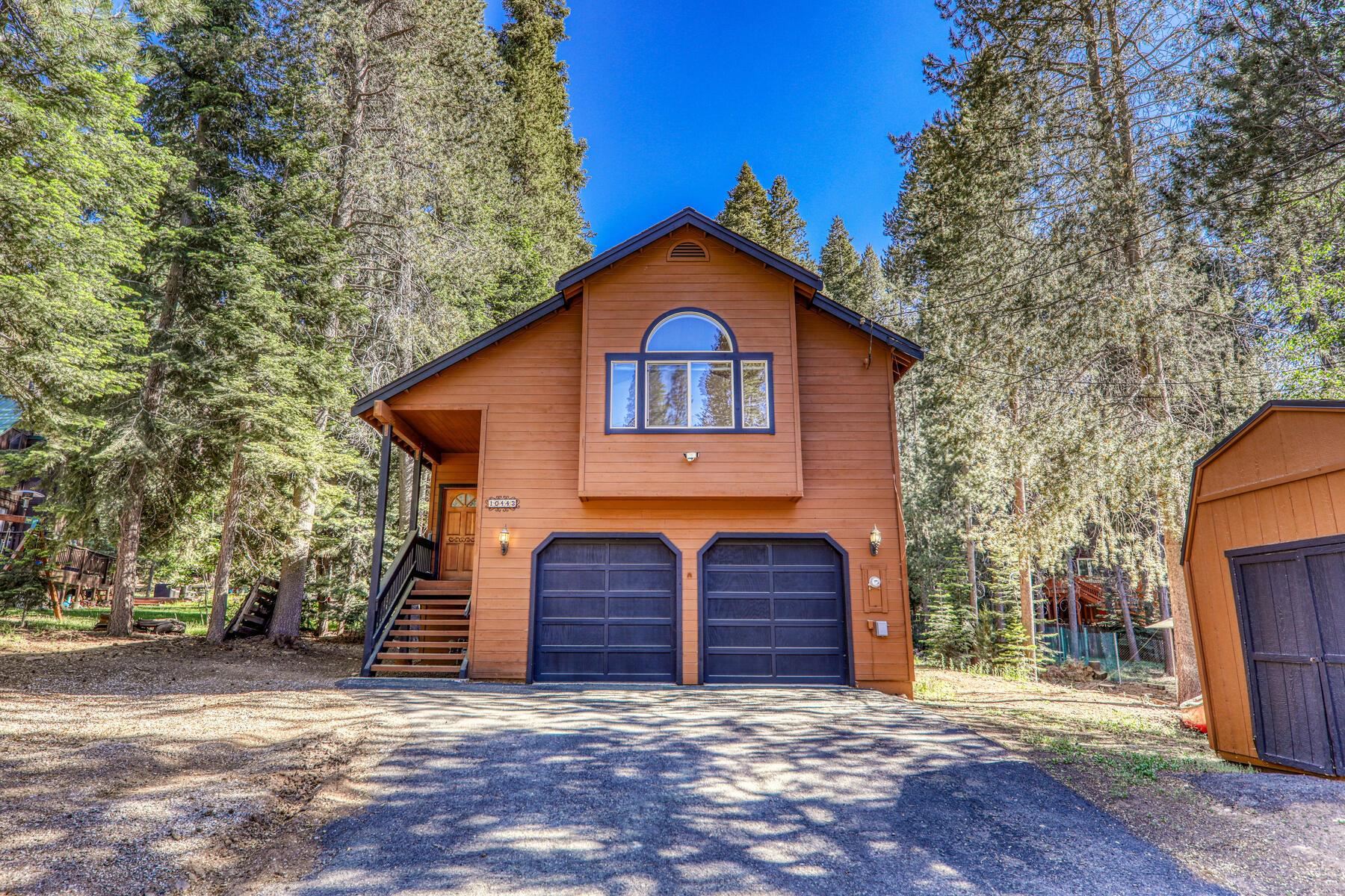 Wood-sided two-story home with double garage and a small outbuilding among tall evergreen trees.
