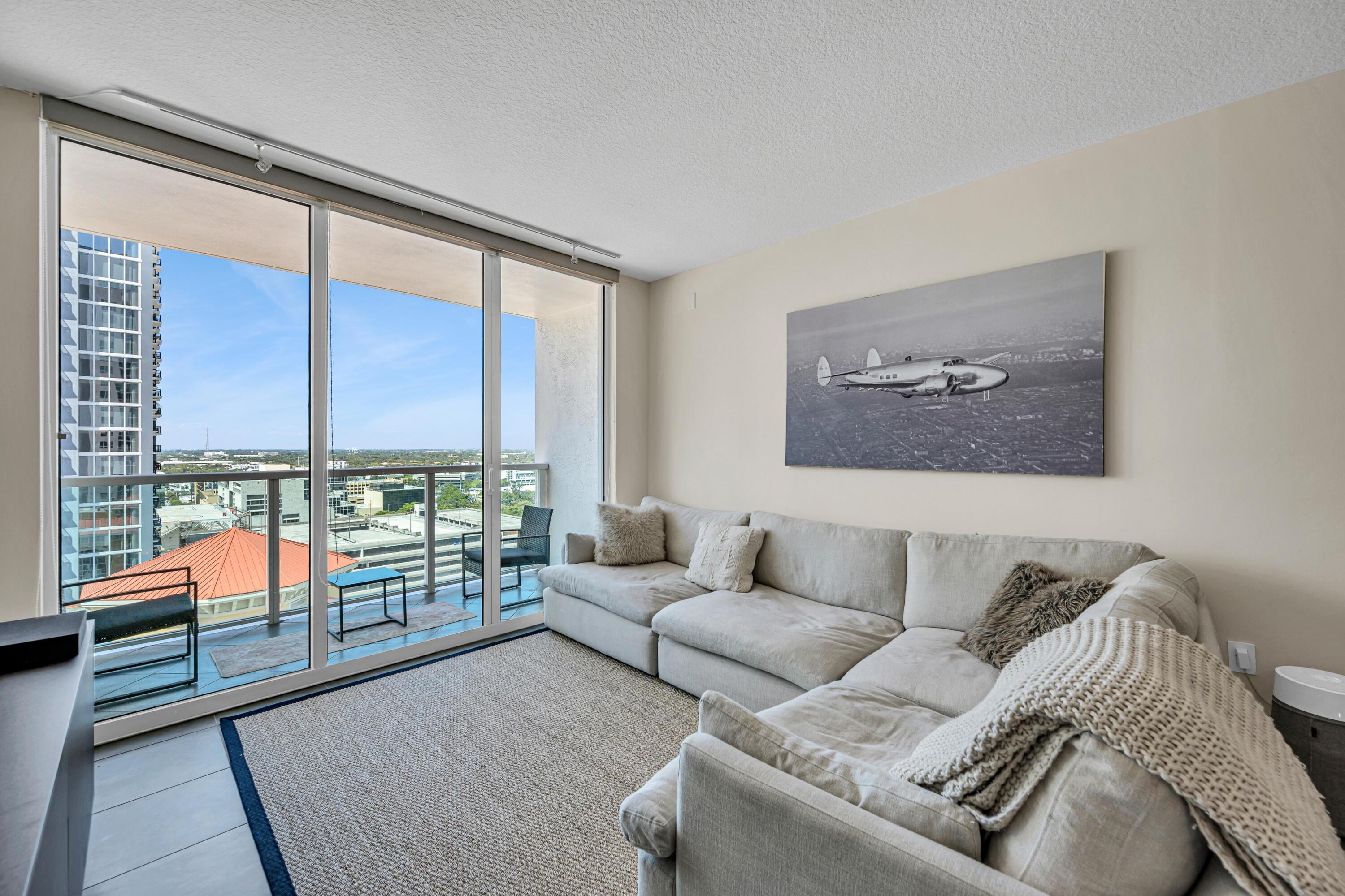 Furnished condo living room with a large sectional, balcony, and urban view through floor-to-ceiling windows.