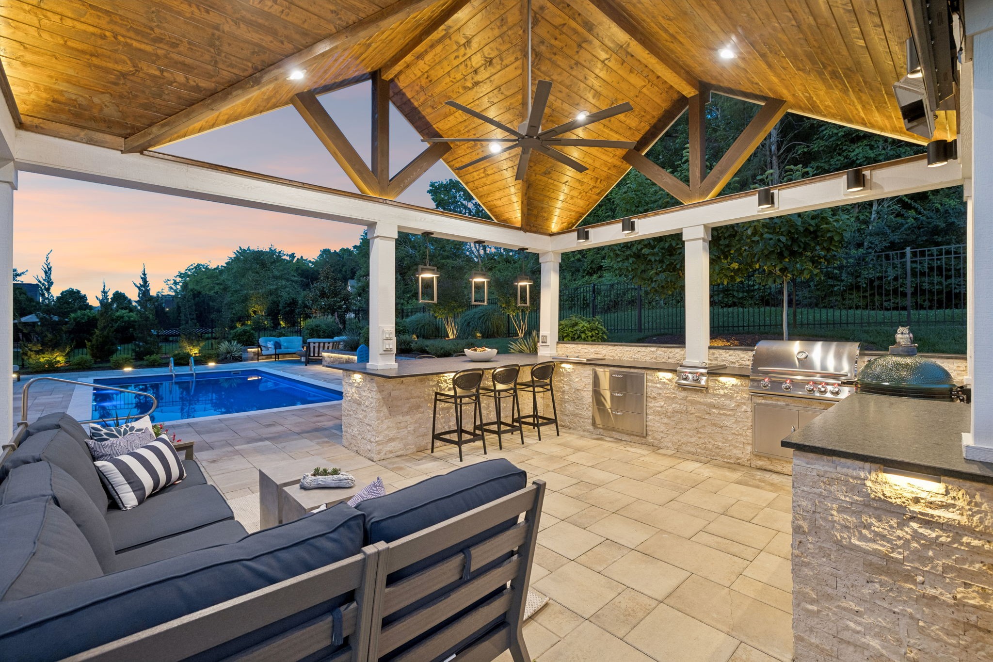 Covered patio with timber ceiling, outdoor kitchen, lounge seating, and swimming pool at dusk.