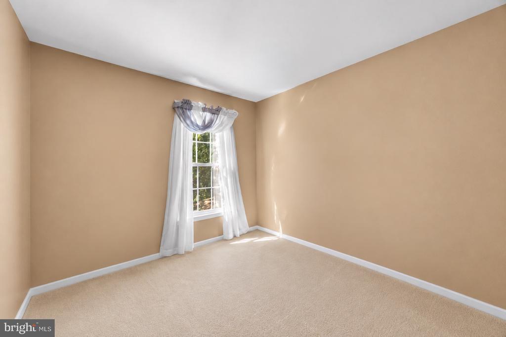 Empty carpeted bedroom with tan walls and a window framed by sheer white curtains.