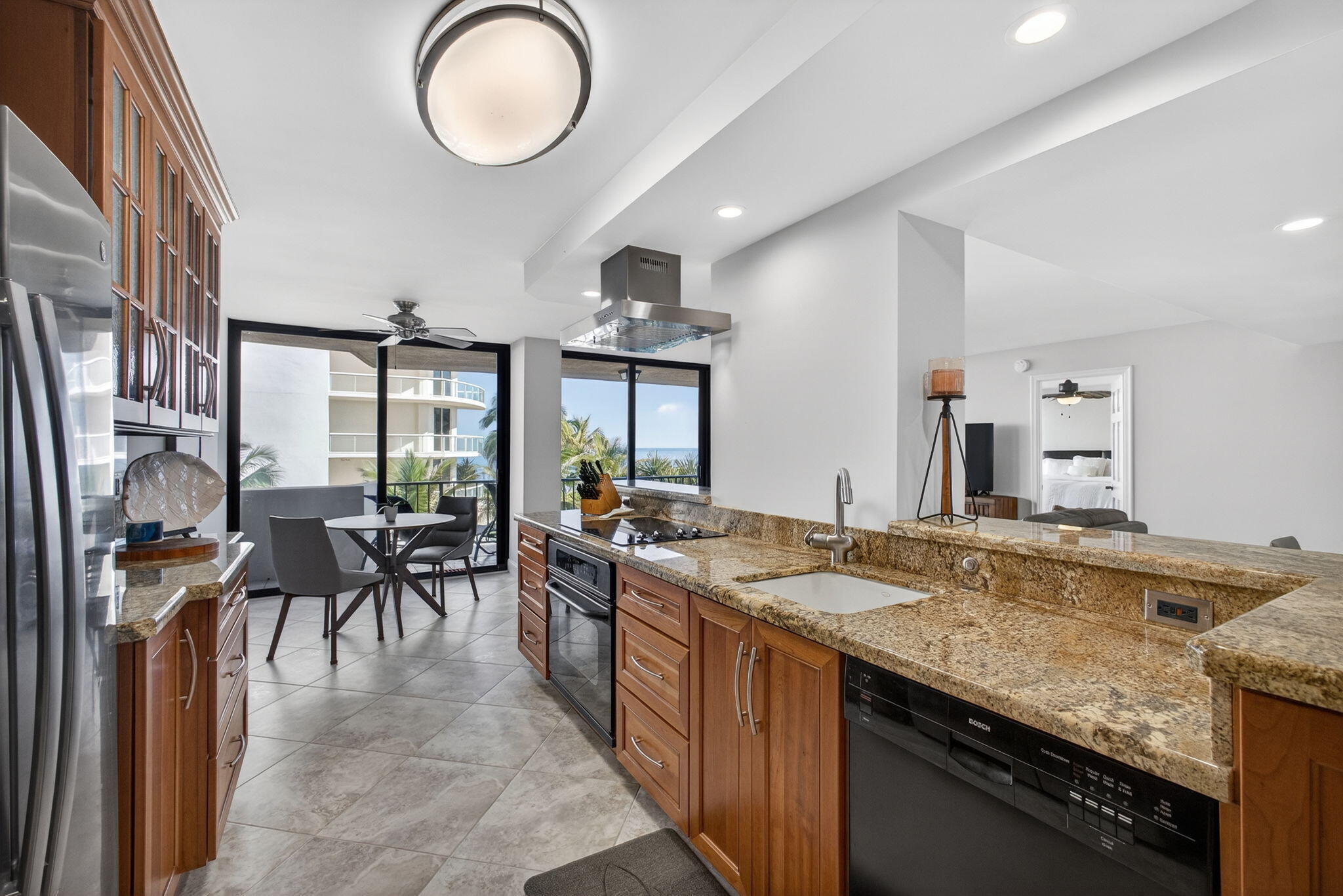 Open kitchen with granite island, warm wood cabinets, and windows overlooking palms and water.