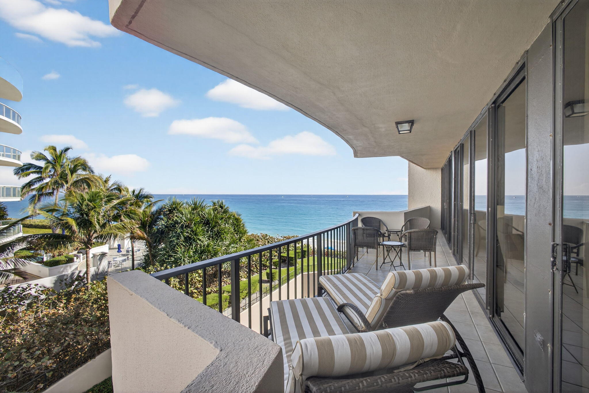 Furnished ocean-view balcony with striped chaise lounges and seating overlooking palm trees and blue water.