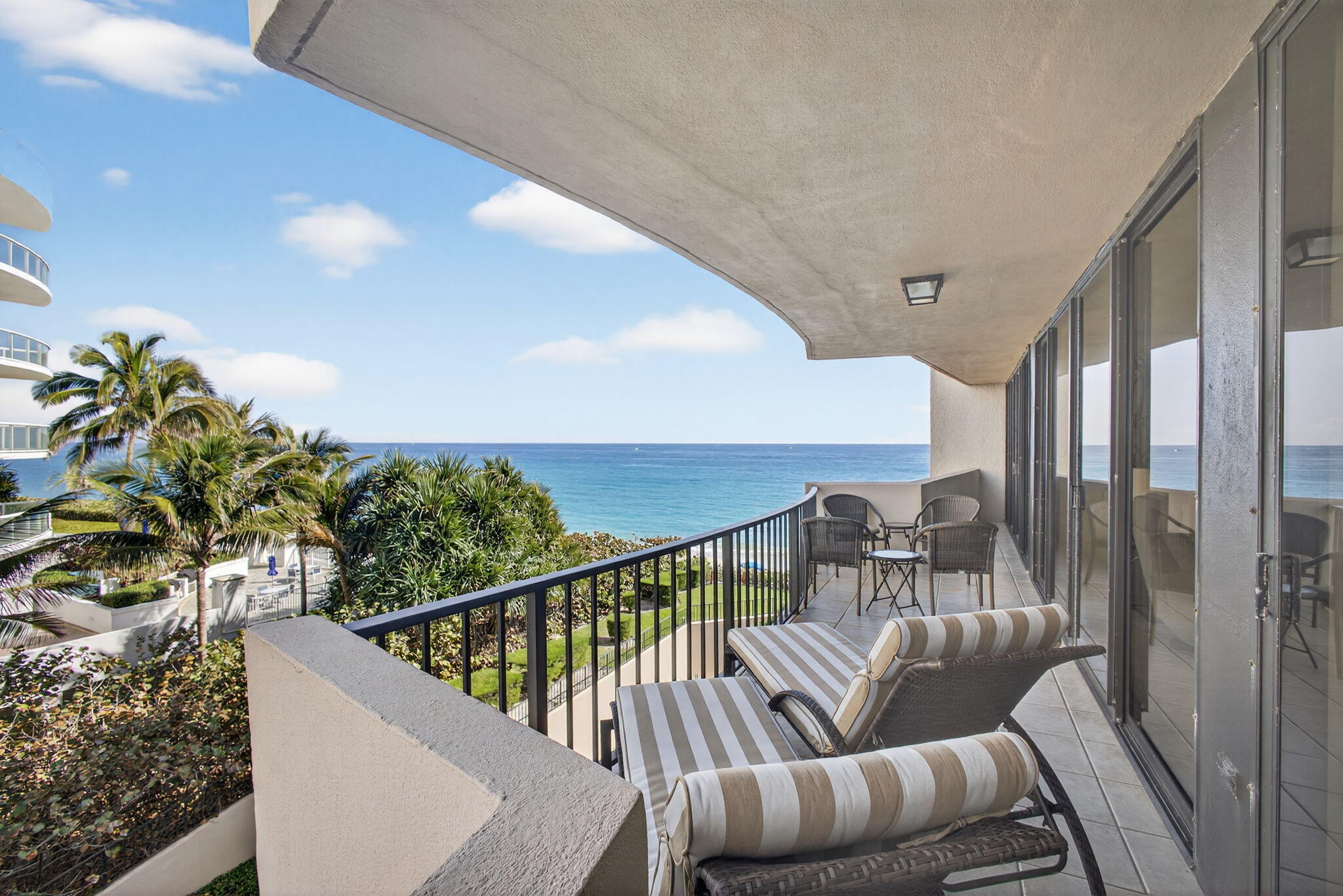 Furnished ocean-view balcony with striped loungers, patio seating, and palm trees below.
