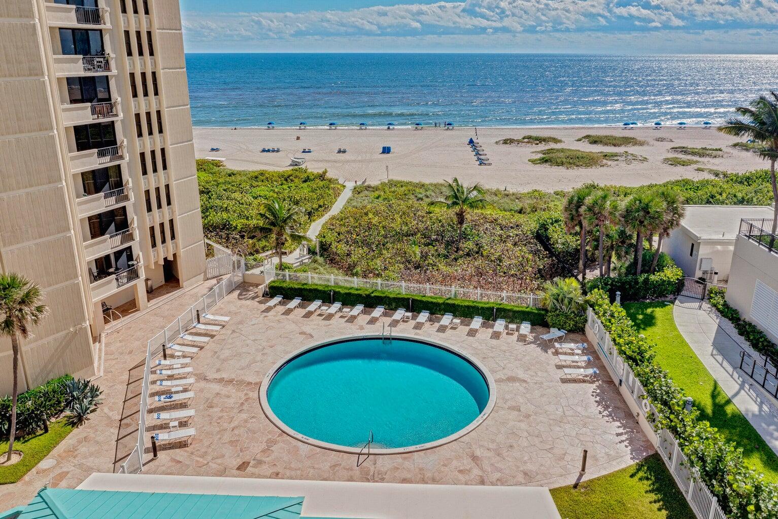 Condominium pool deck overlooking a sandy beach and ocean, with lounge chairs around the rounded pool.