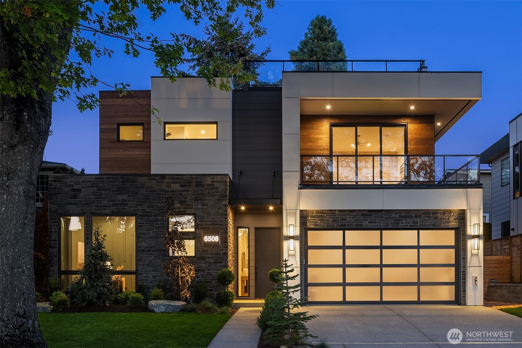 Modern two-story home at dusk with a stone facade, illuminated garage, and glass-railed balcony.