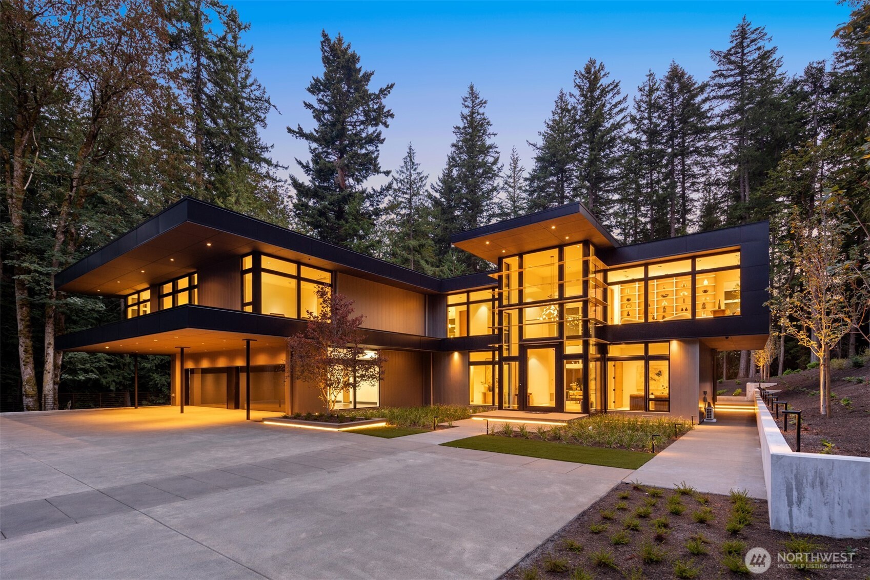 Modern two-story home with illuminated glass walls, a broad driveway, and evergreen trees at dusk.