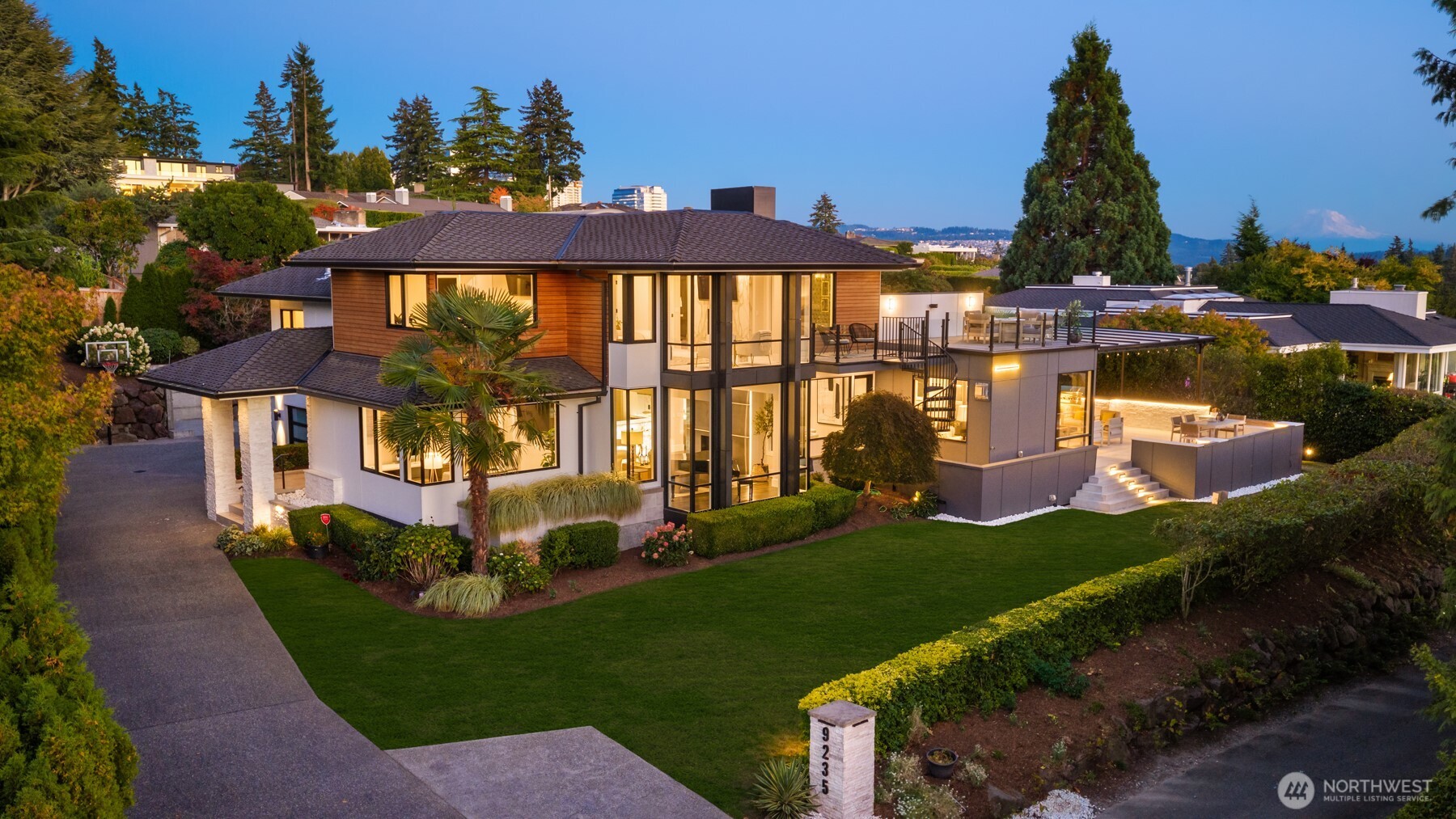 Modern two-story home with expansive windows, landscaped lawn, and illuminated terraces at dusk.