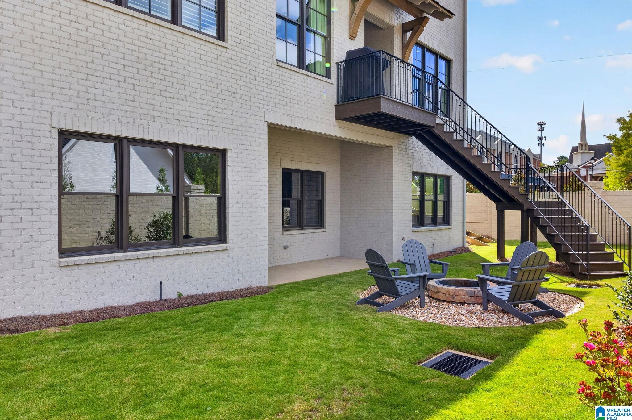 White brick home’s backyard with an exterior staircase, balcony, and fire pit surrounded by Adirondack chairs.