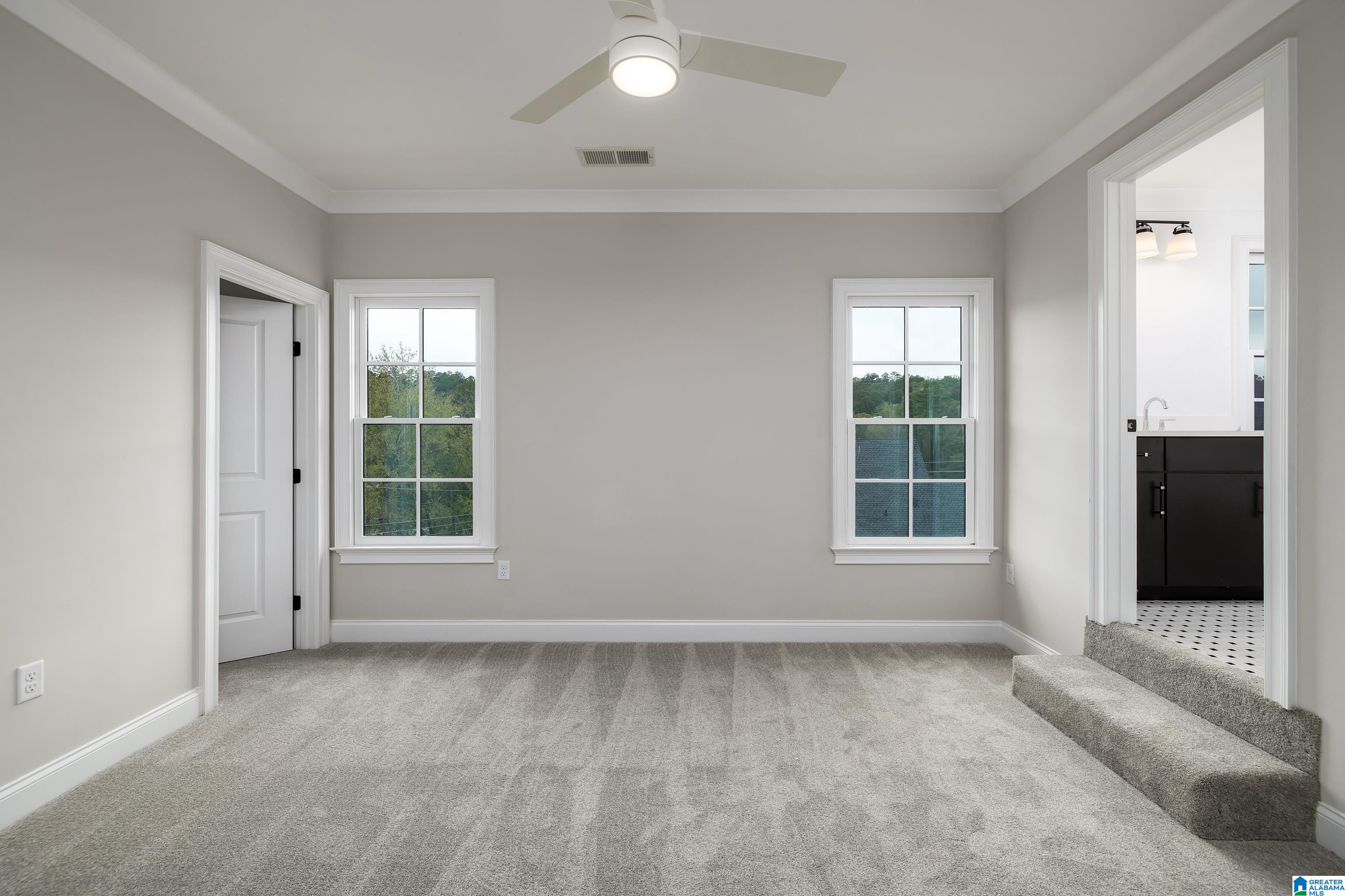 Empty carpeted bedroom with two windows, a ceiling fan, and an adjoining bathroom.
