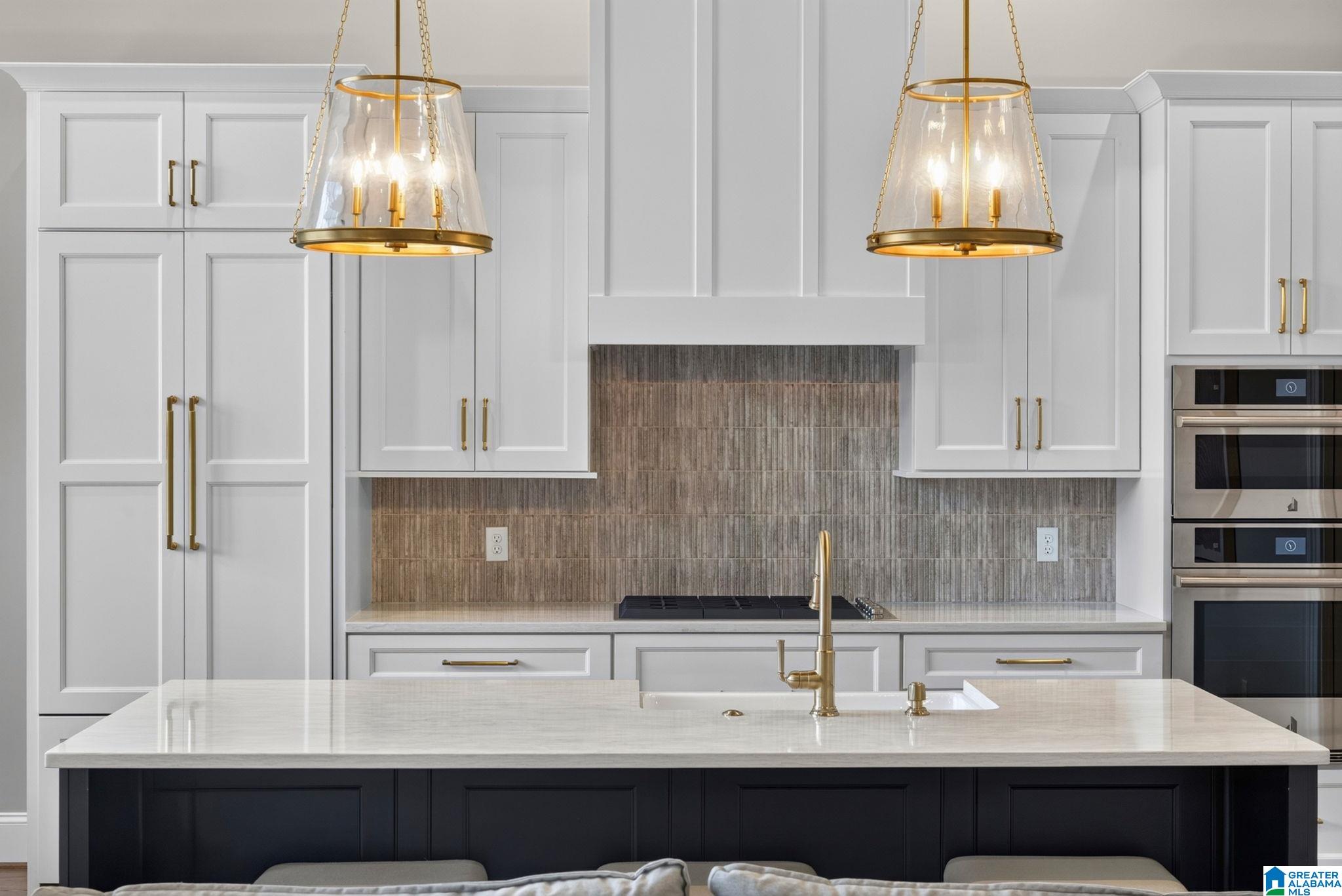 Modern kitchen with a dark island, white cabinetry, textured backsplash, brass pendants, and double ovens.