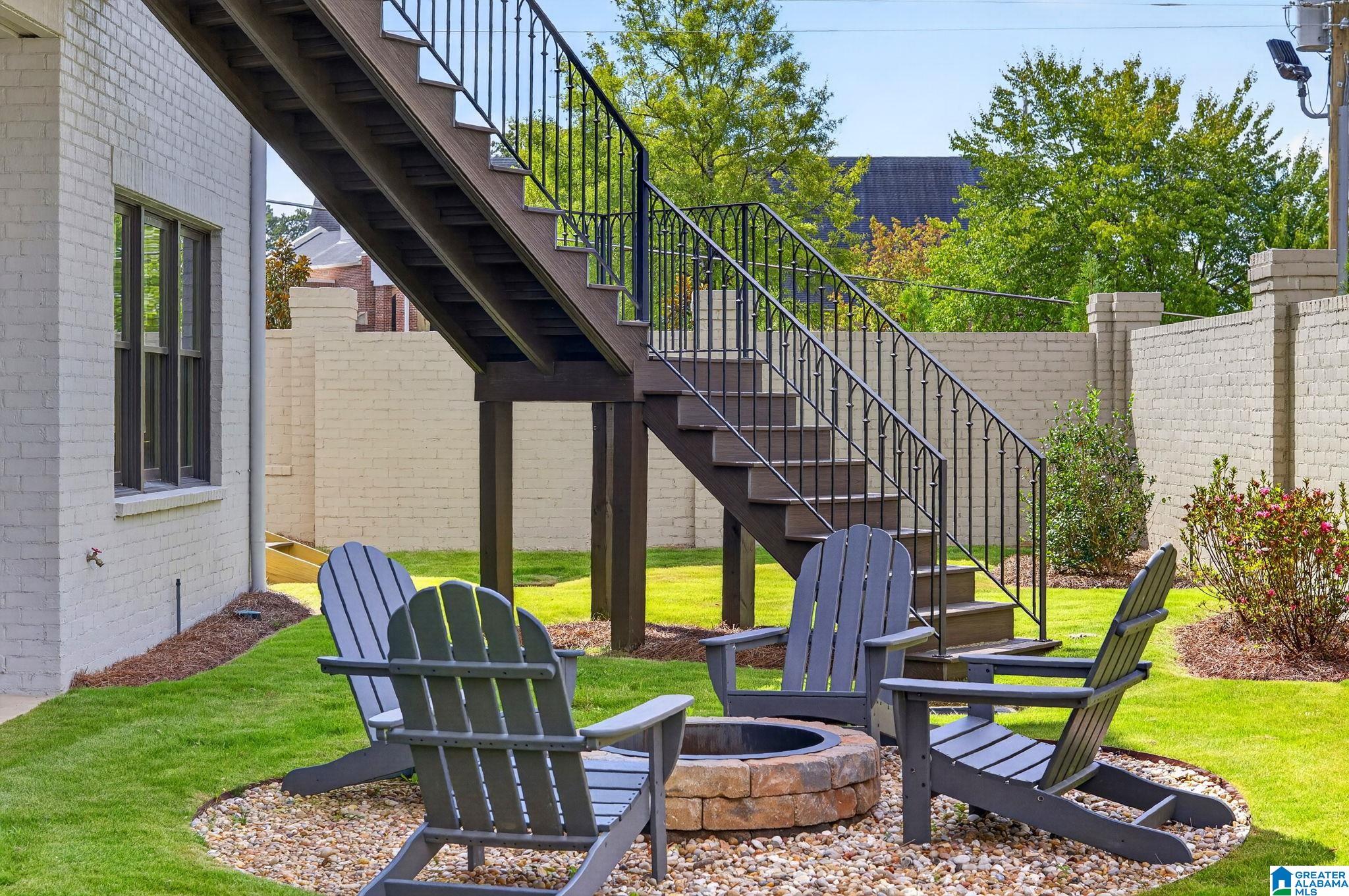 Backyard patio with Adirondack chairs around a stone fire pit beneath an elevated staircase.