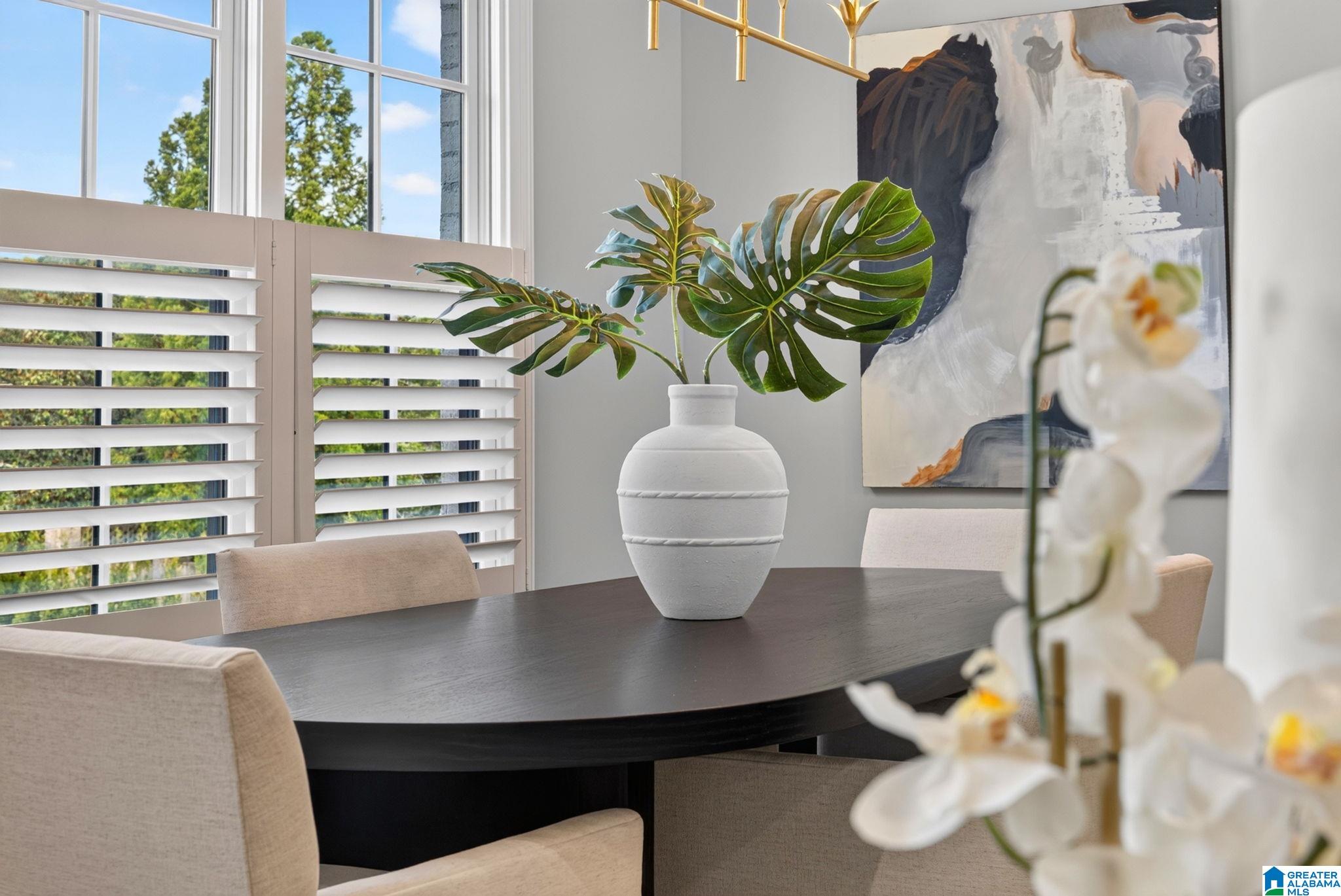 Dining room with a dark oval table, plantation shutters, and a leafy arrangement in a white vase.