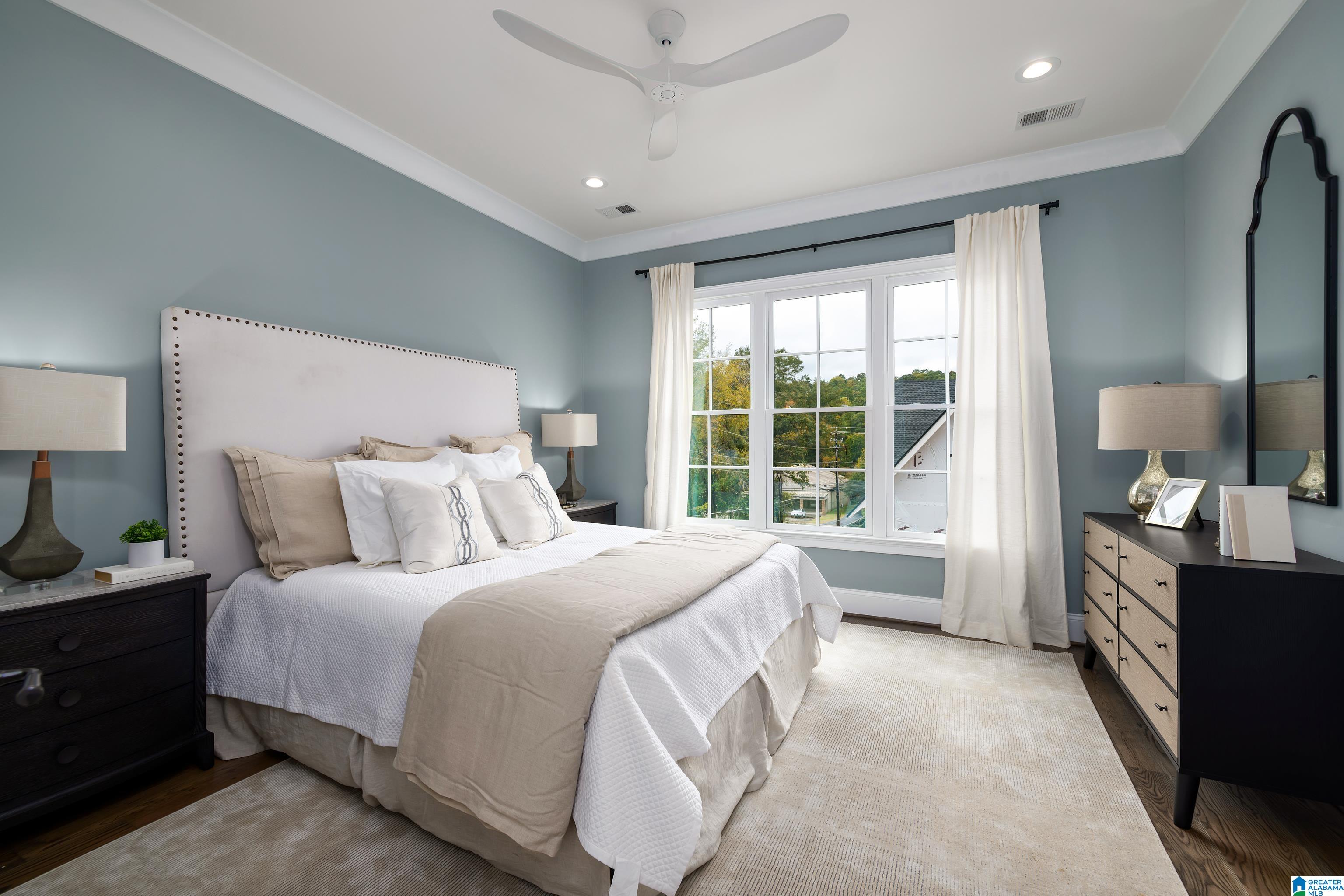 Bedroom with a nailhead-trim headboard, white and beige bedding, and a large curtained window.