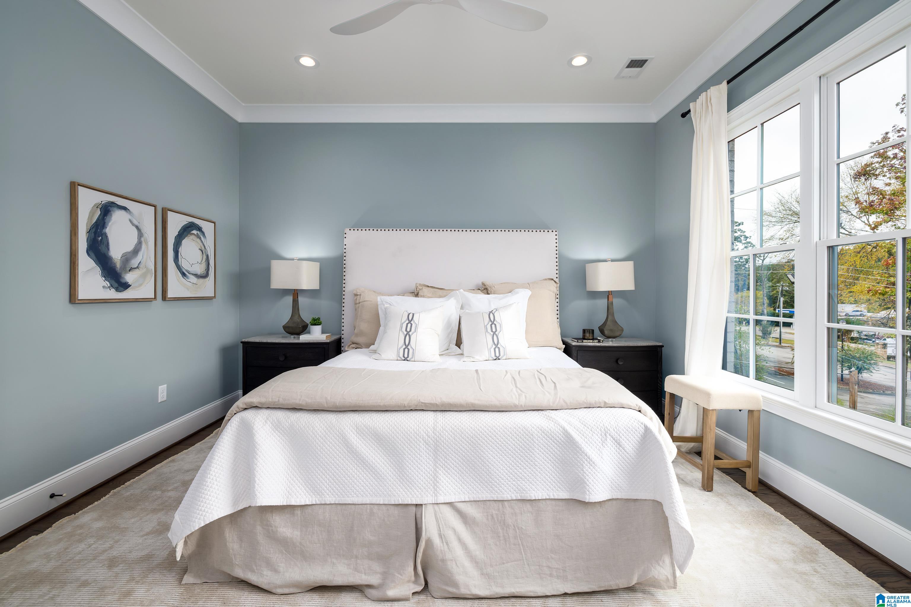 Staged bedroom with a white bed, blue-gray walls, abstract art, and tall windows.