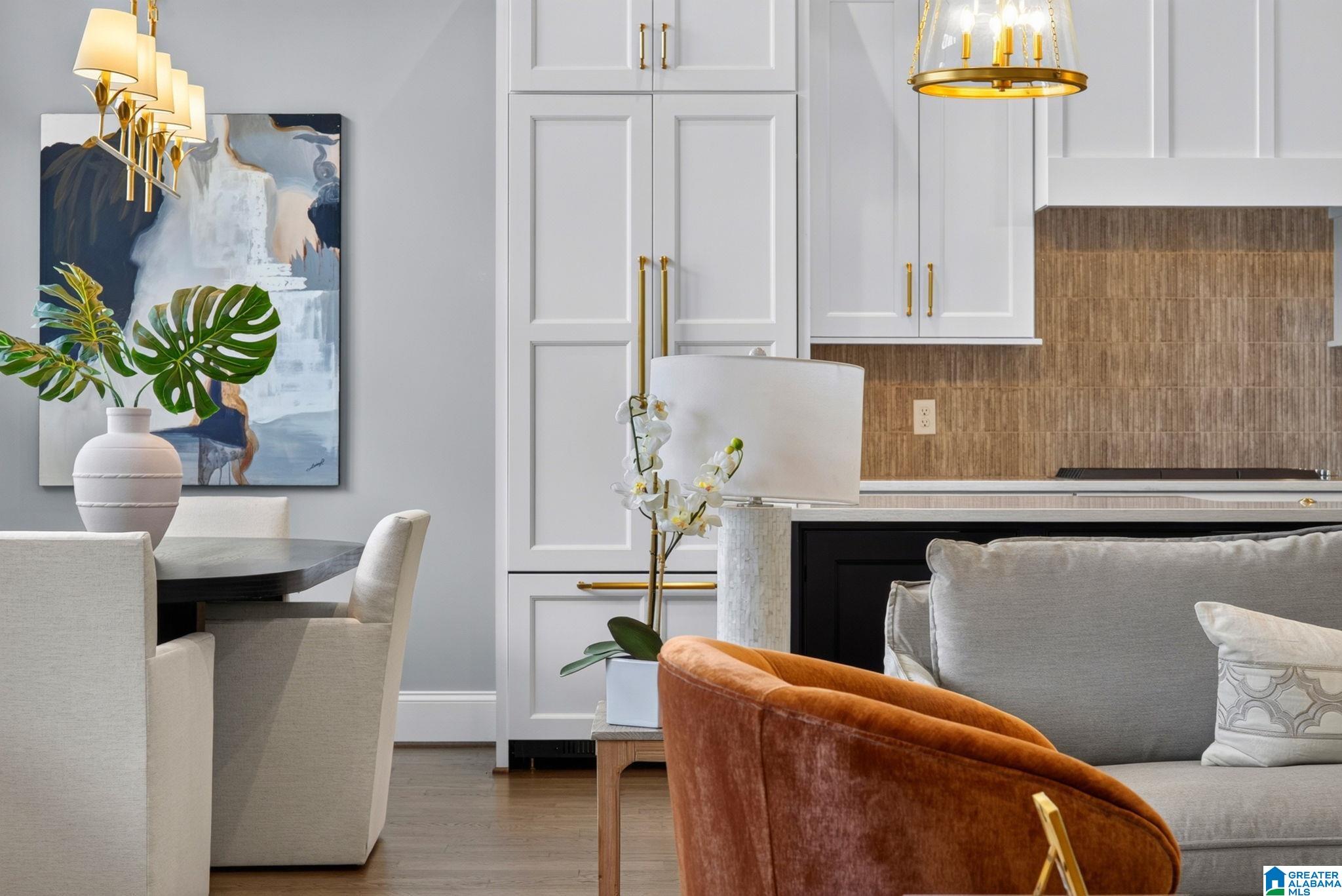 Open-plan kitchen and dining area with white cabinets, brass fixtures, textured backsplash, and colorful furnishings.