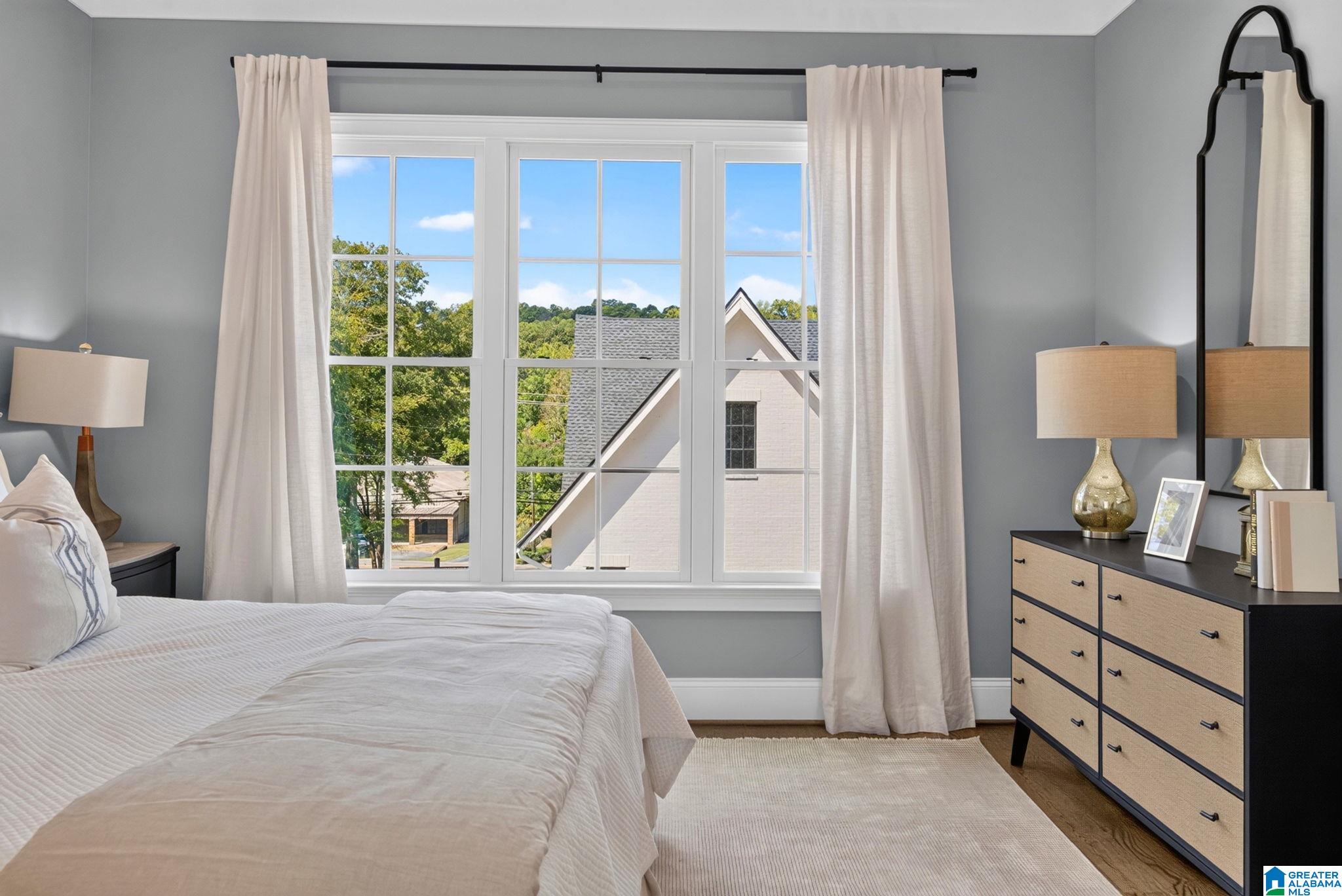 Staged bedroom with a white bed, large curtained window, and dresser with a tall mirror.