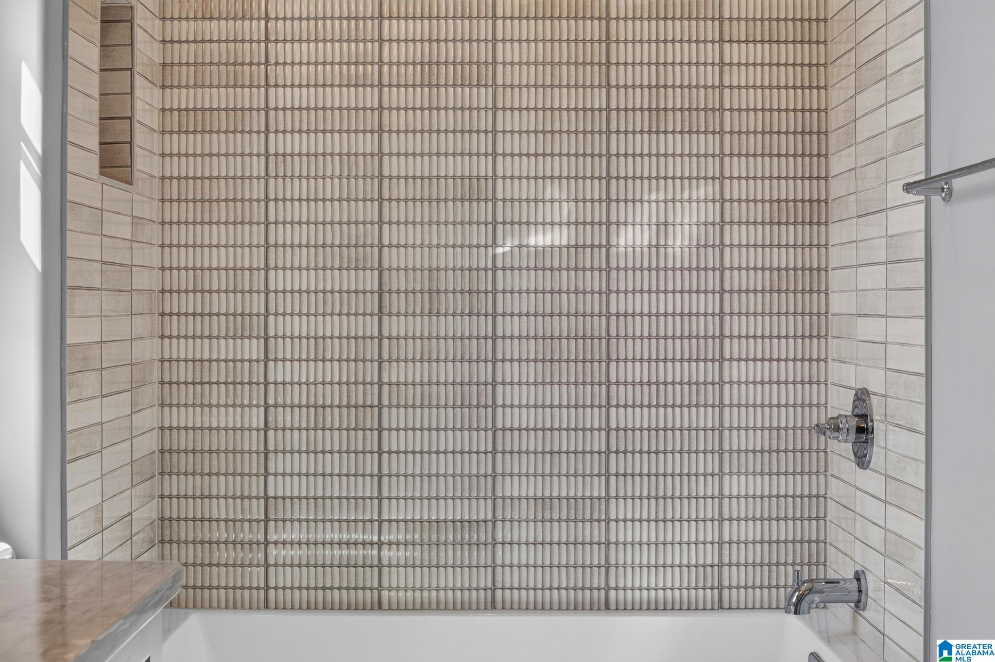Bathroom tub and shower with textured beige tile, chrome fixtures, and a stone vanity edge.