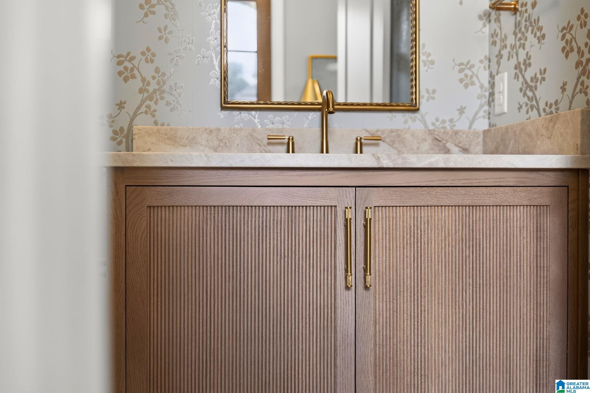 Bathroom vanity with fluted wood cabinetry, marble counter, brass fixtures, and floral wallpaper.