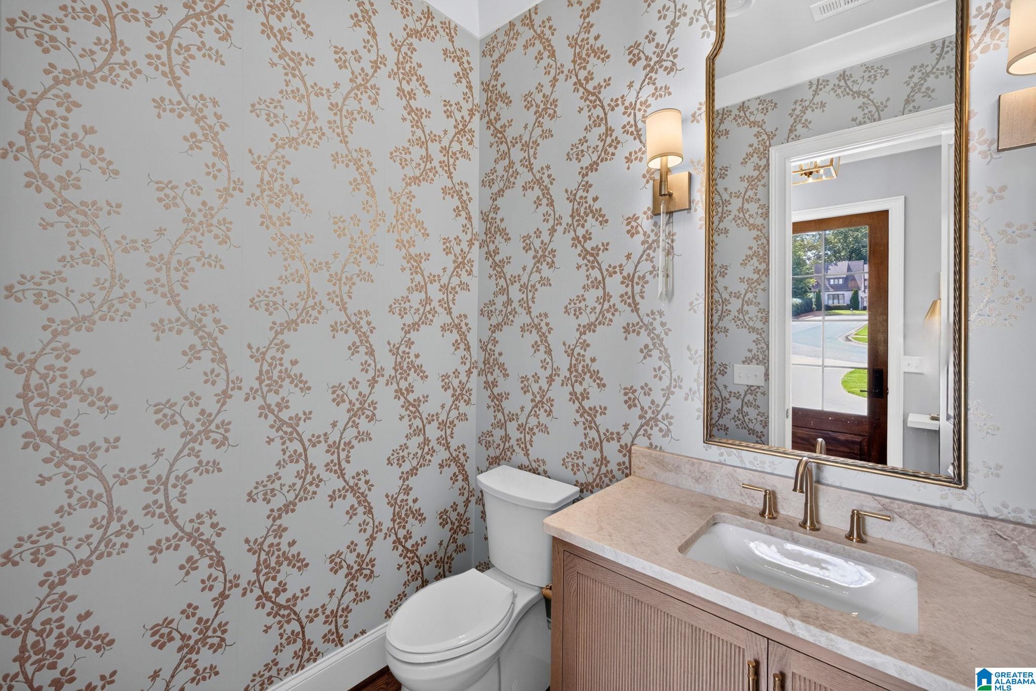 Powder room with botanical wallpaper, a wood vanity, pale stone countertop, and large framed mirror.