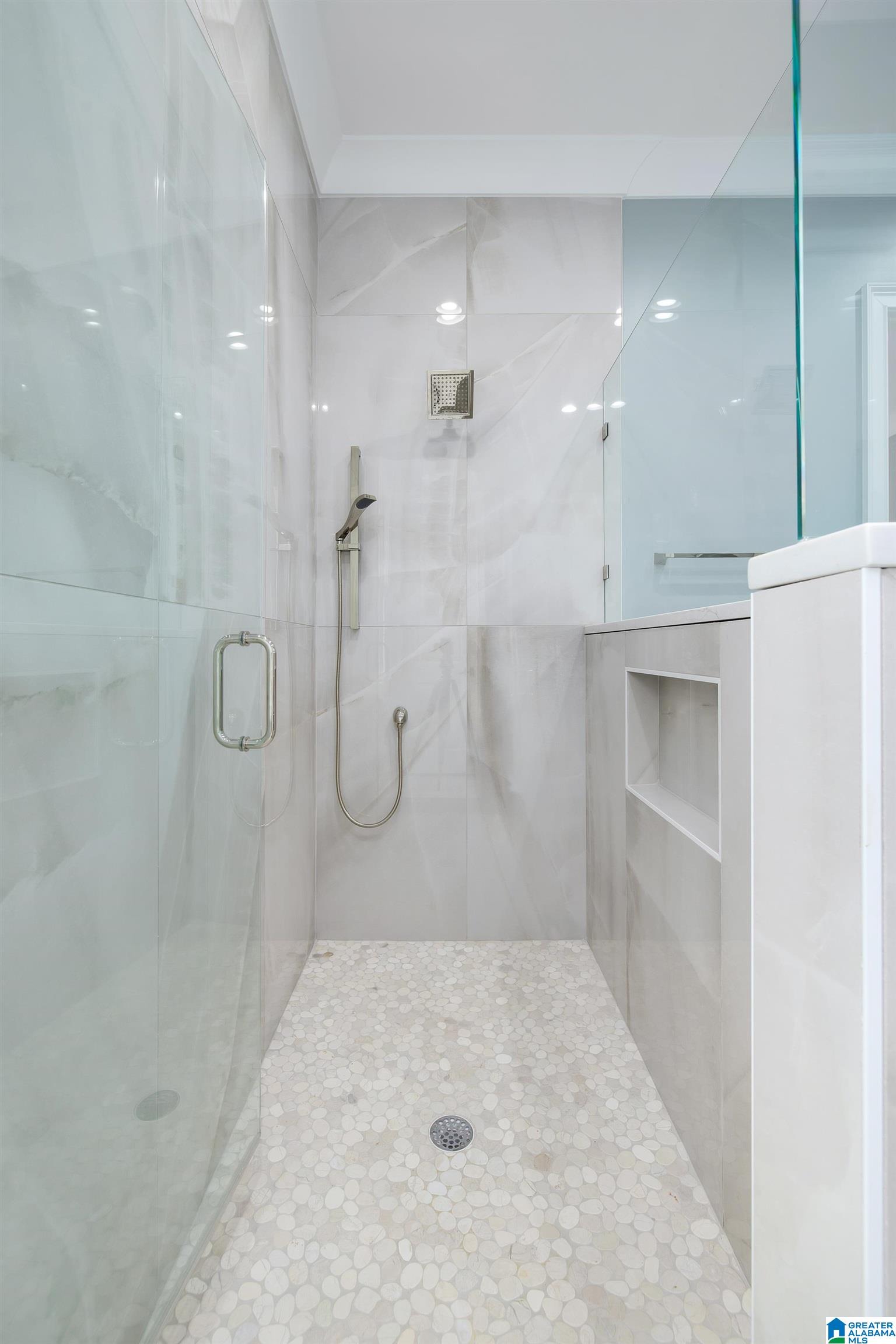 Walk-in shower with glass panels, marble-look tile, a handheld showerhead, and pebble-mosaic flooring.