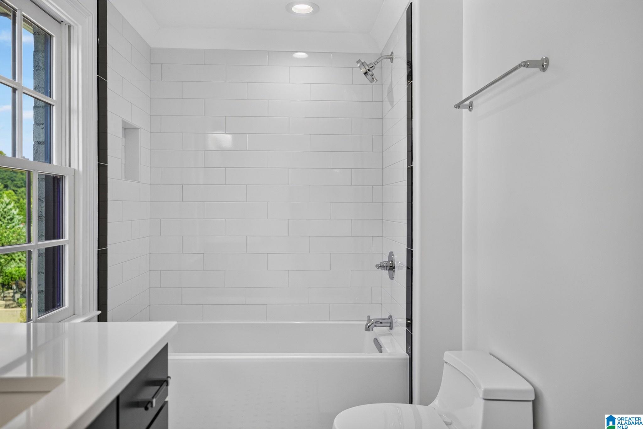 Bathroom with a white tiled tub and shower, large window, vanity, and toilet.