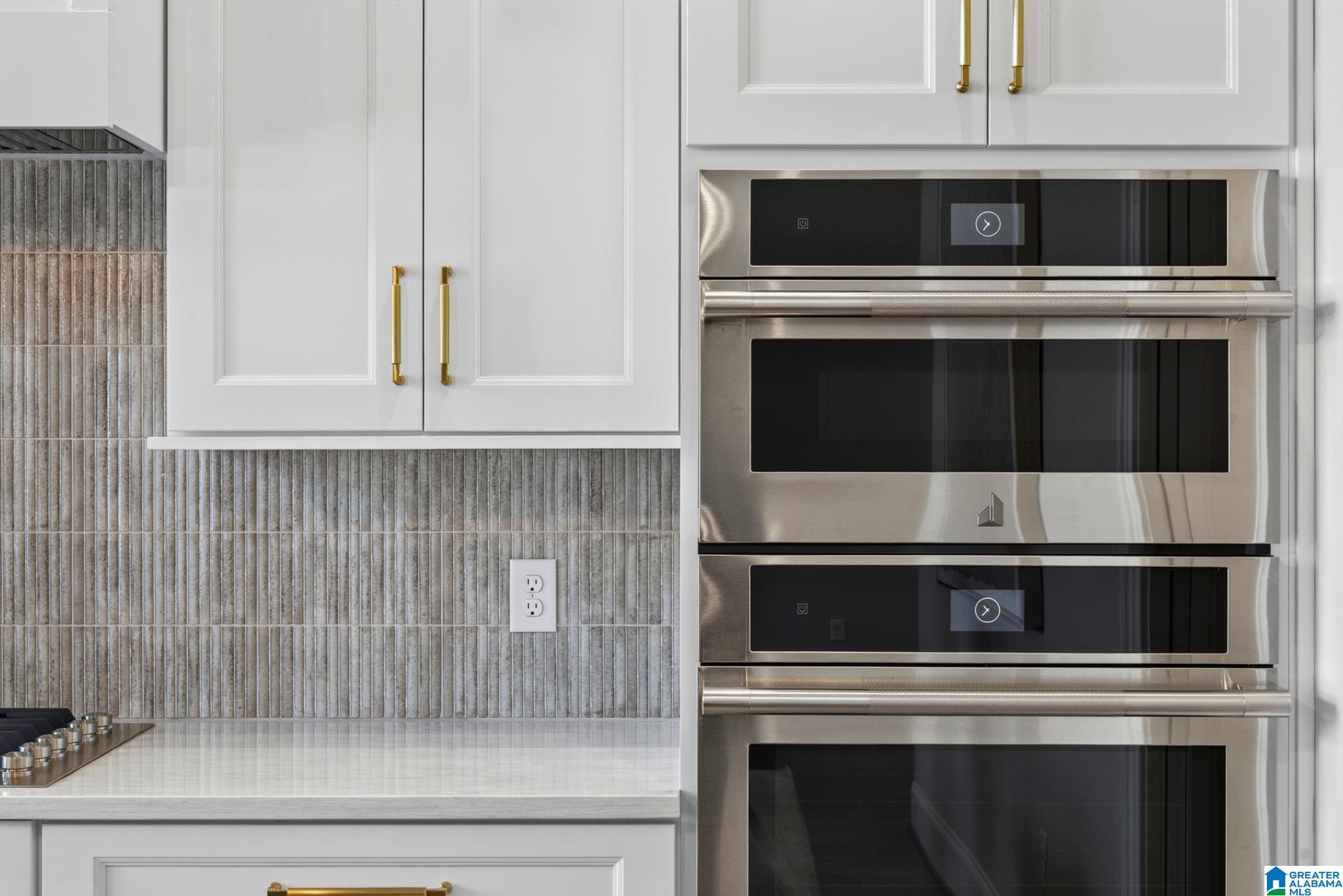 Kitchen with stacked stainless-steel ovens, white cabinets, brass pulls, and a gray tile backsplash.
