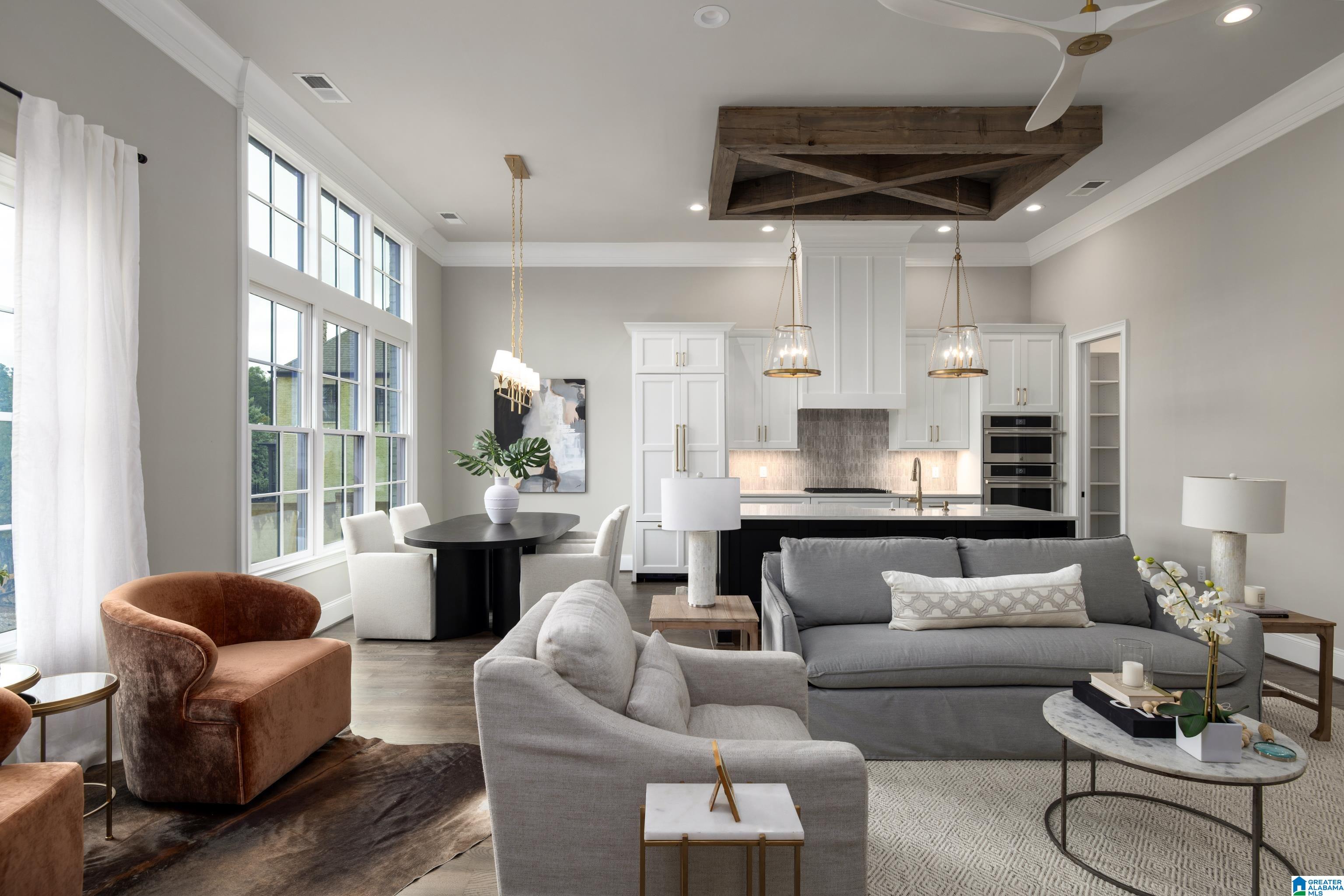 Open-plan living room and kitchen with tall windows, gray sofas, and a rustic wood ceiling feature.