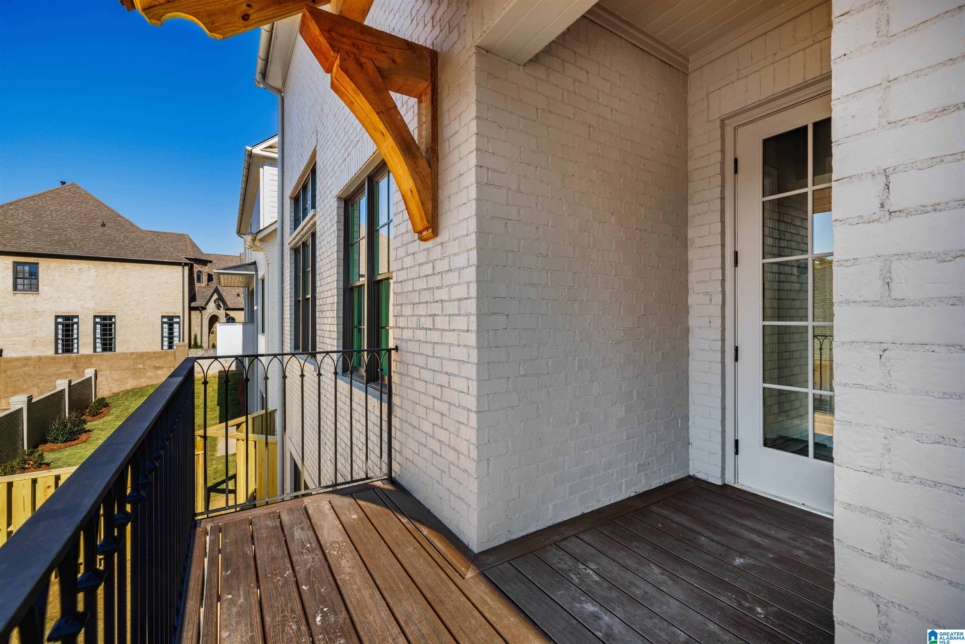 Raised wooden balcony with black railing on a white brick home, overlooking a fenced lawn and neighboring house.