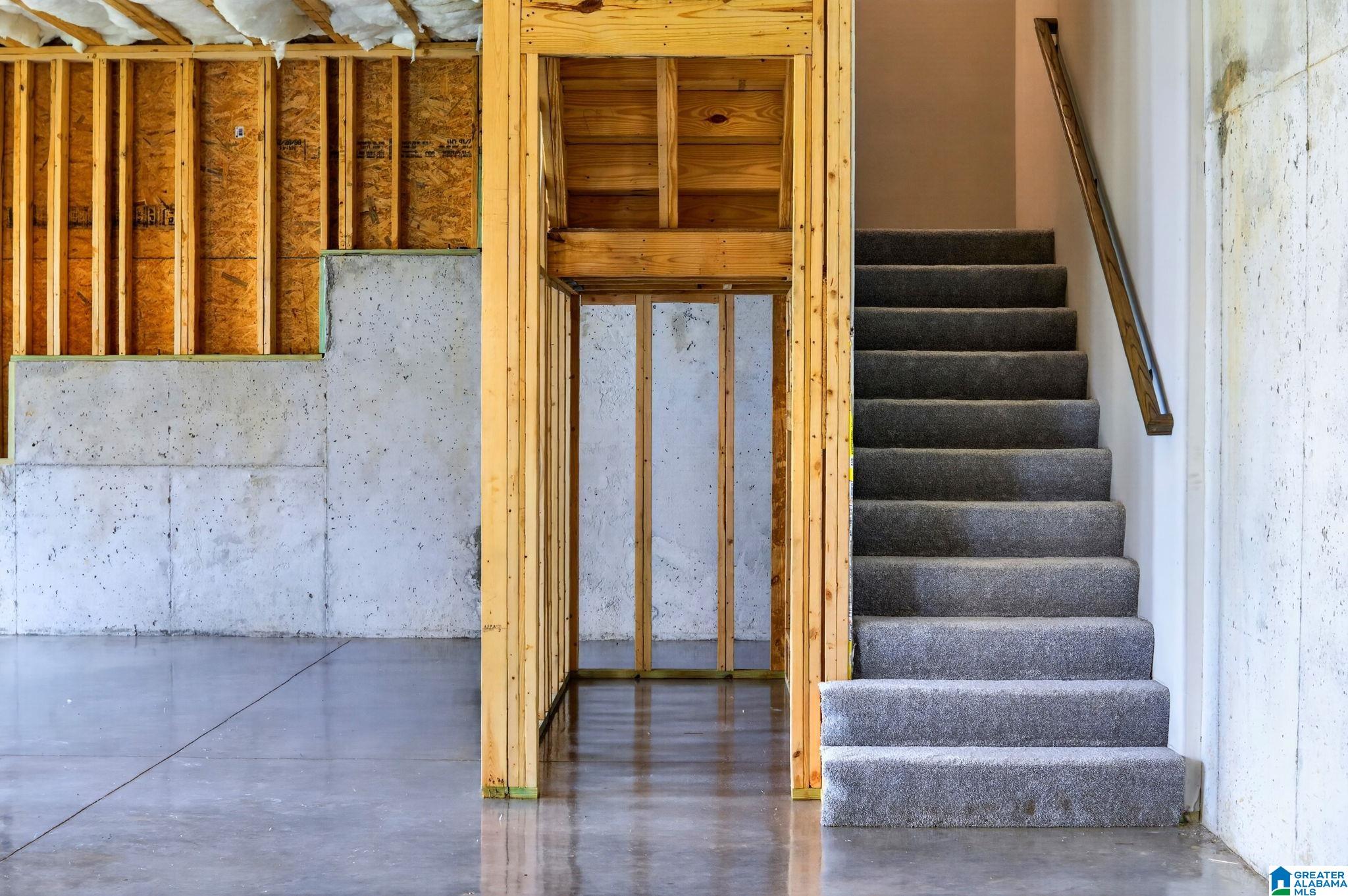 Unfinished basement with exposed framing, concrete walls and floor, and carpeted stairs.