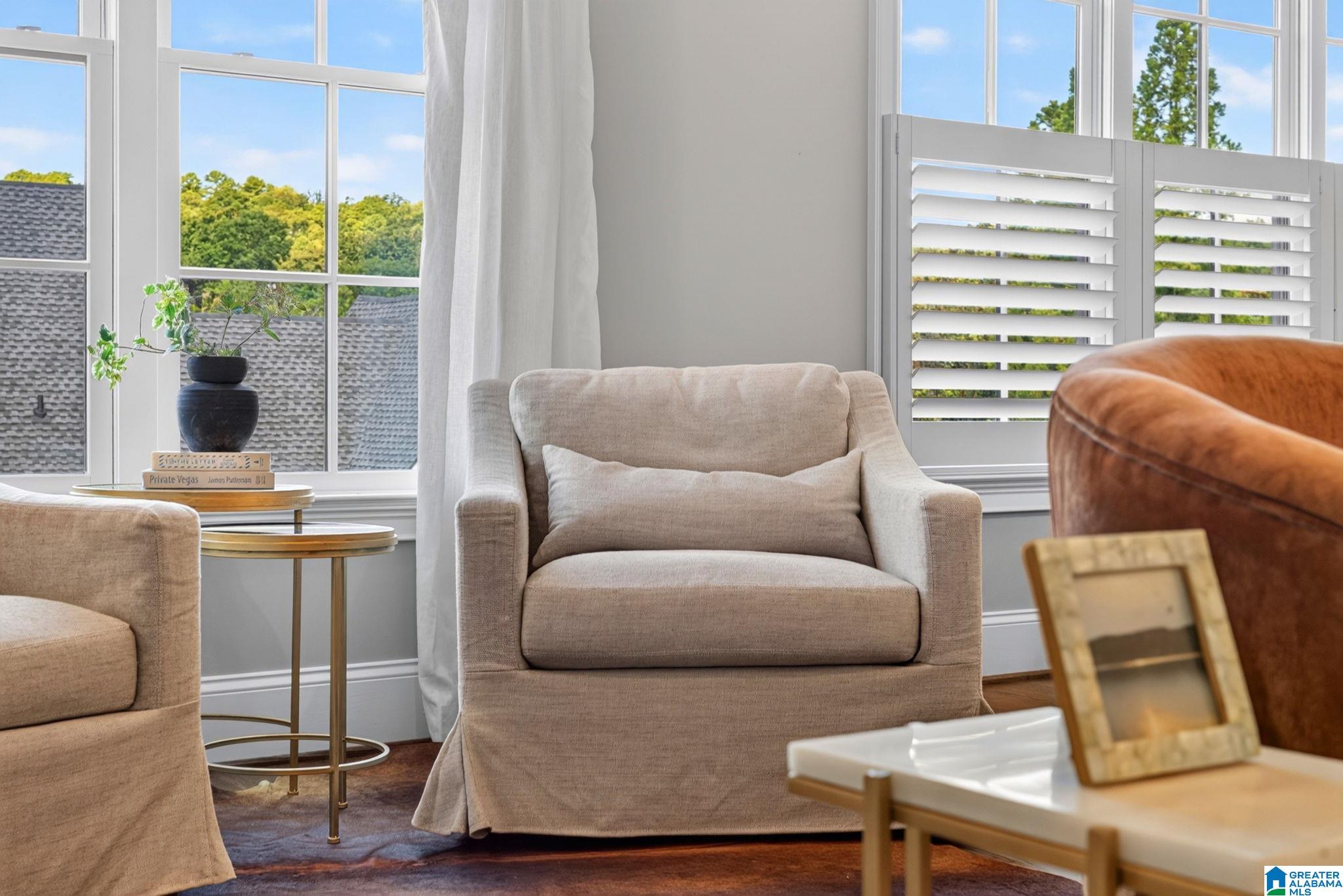 Furnished living room with a beige armchair, bright windows, and white plantation shutters.