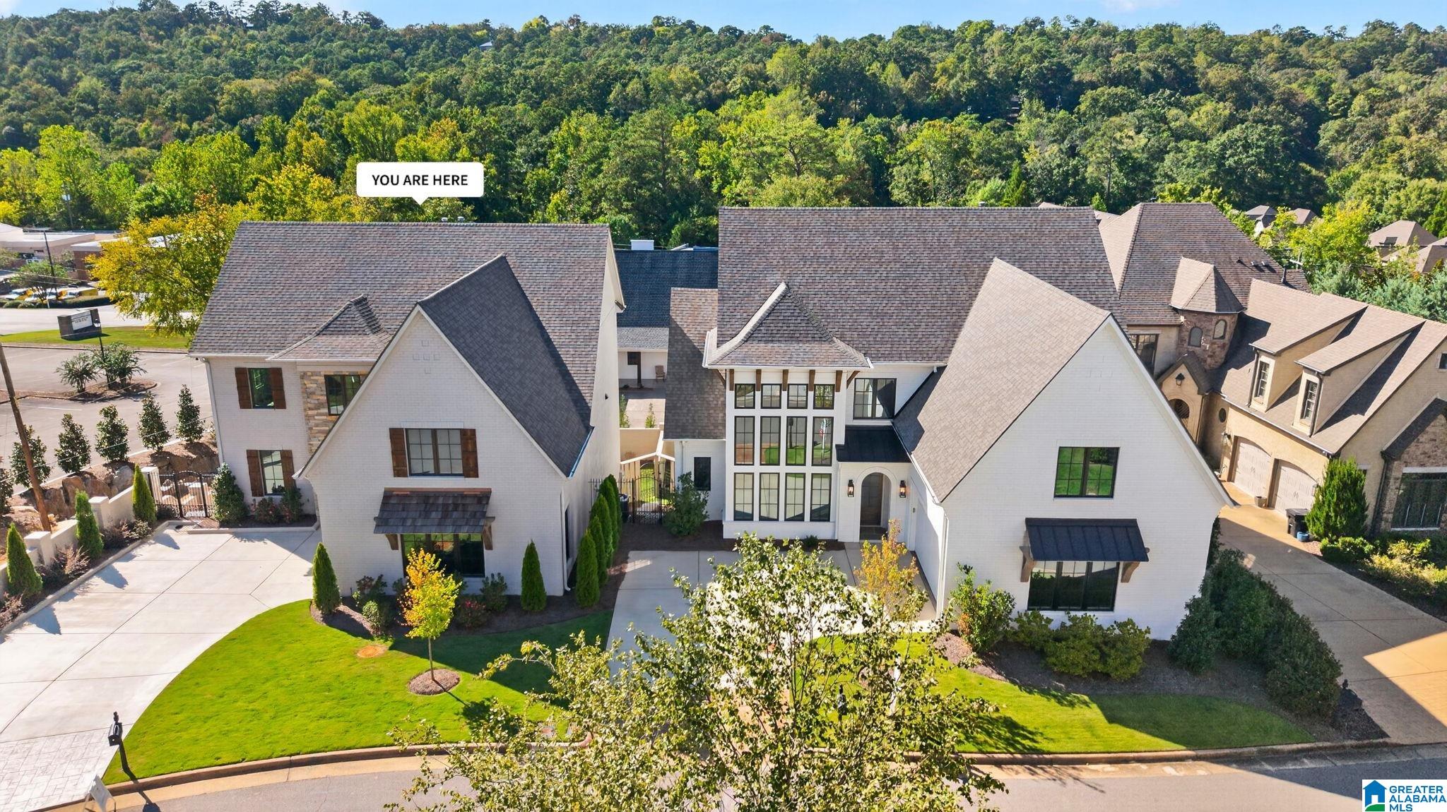 Aerial view of a large white-brick home with a landscaped lawn and wooded hills behind it.