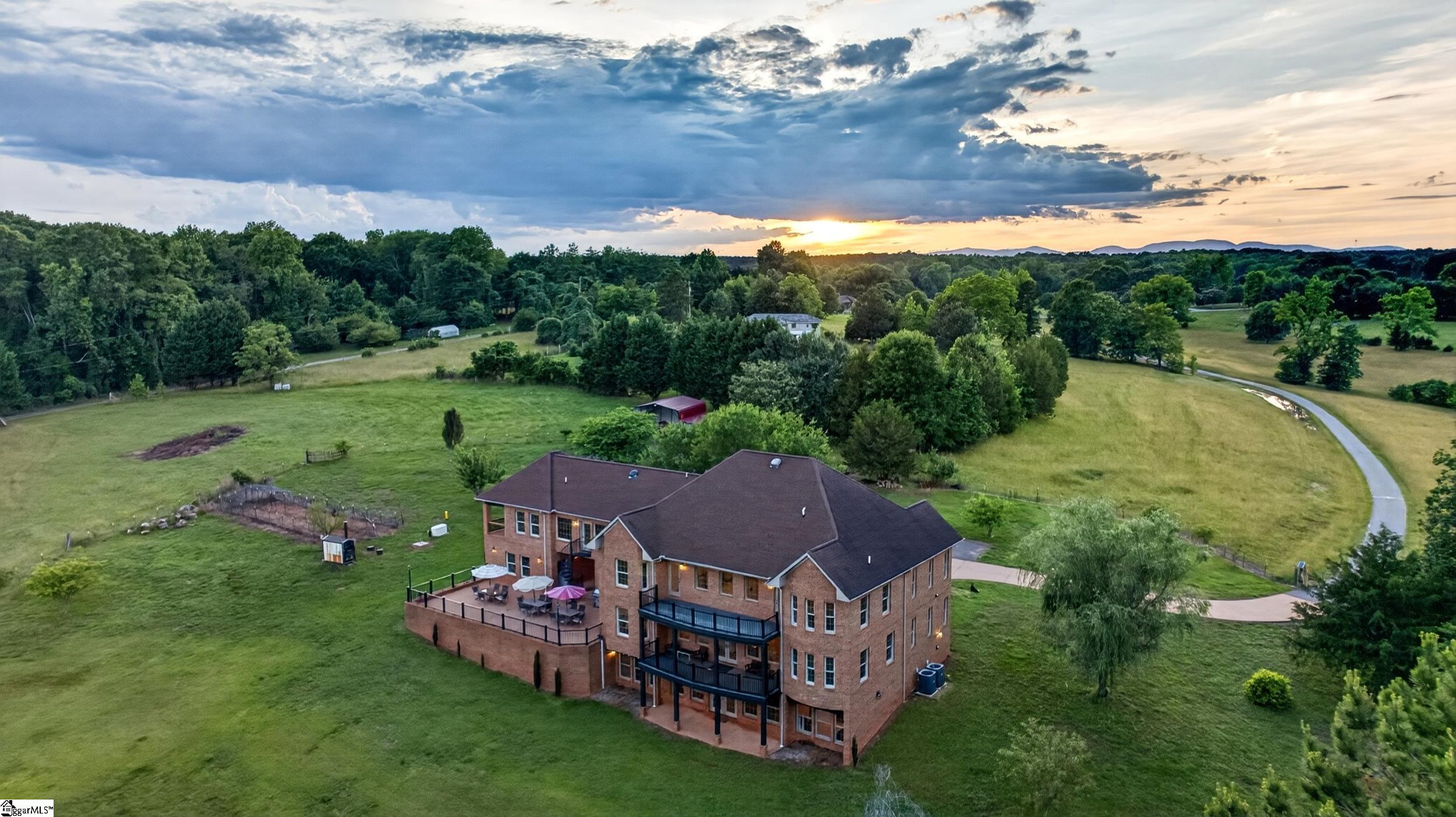 Aerial view of a large brick home surrounded by open lawns and trees, with a fenced patio and distant hills.