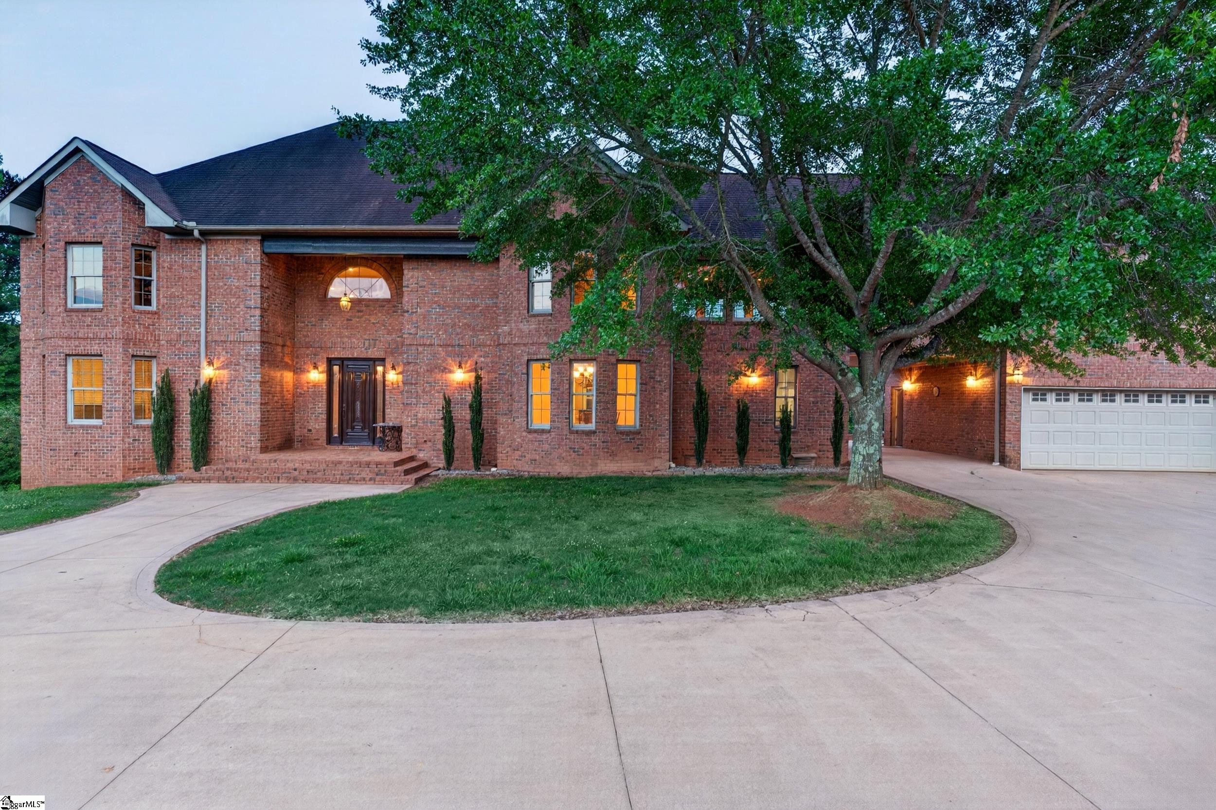 Two-story brick house with a curved driveway, attached garage, and large shade tree at dusk.