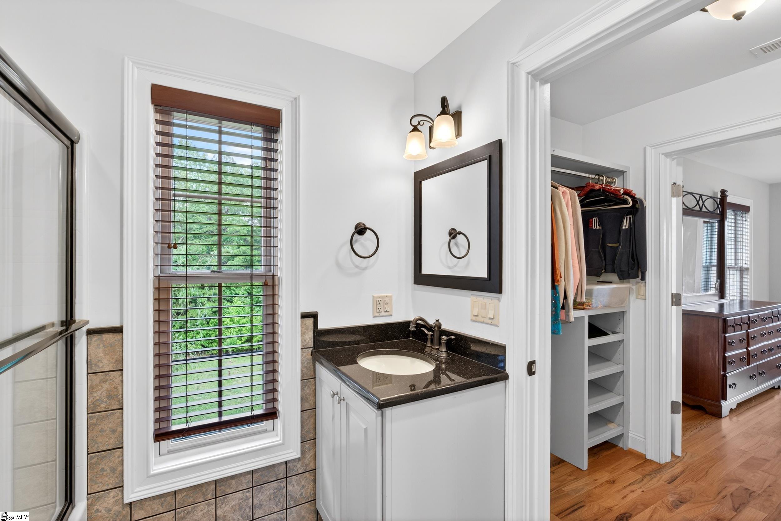 Bathroom vanity with dark stone counter and window beside a glass shower; open closet and bedroom visible beyond.