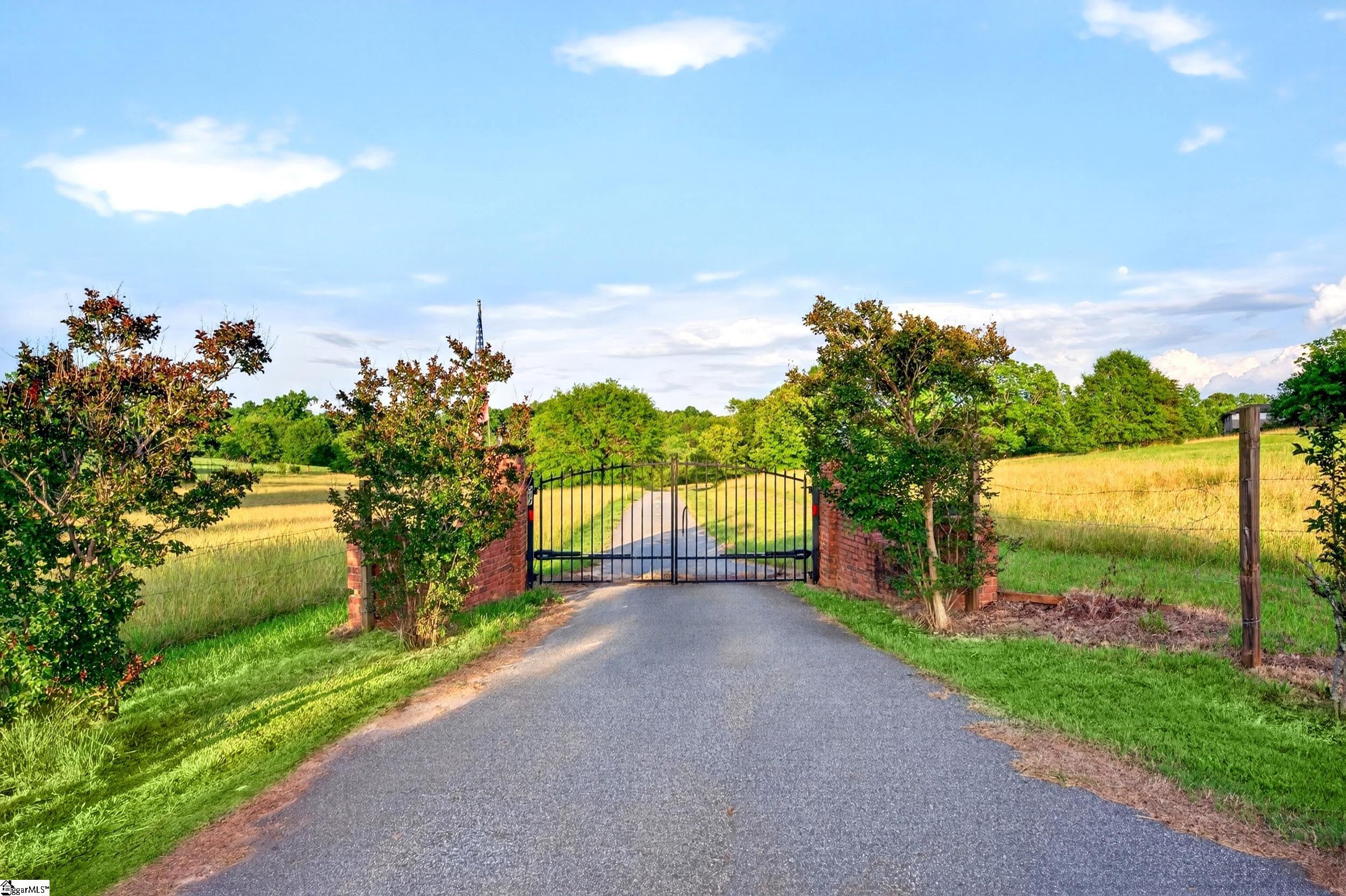 Gated rural driveway with brick pillars, golden fields, and green trees.