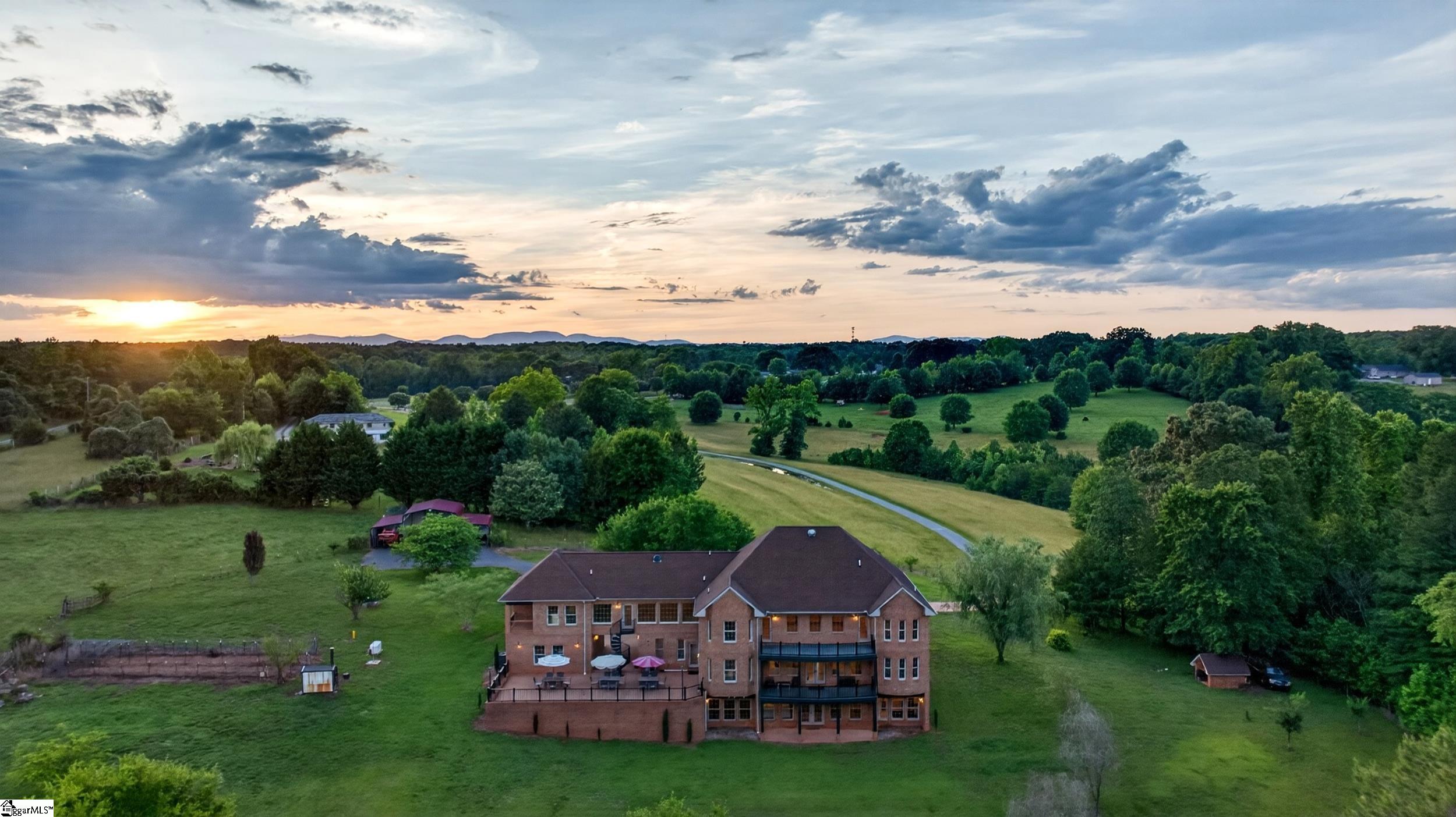 Aerial view of a large brick home on open acreage, with a curved drive and distant hills at sunset.