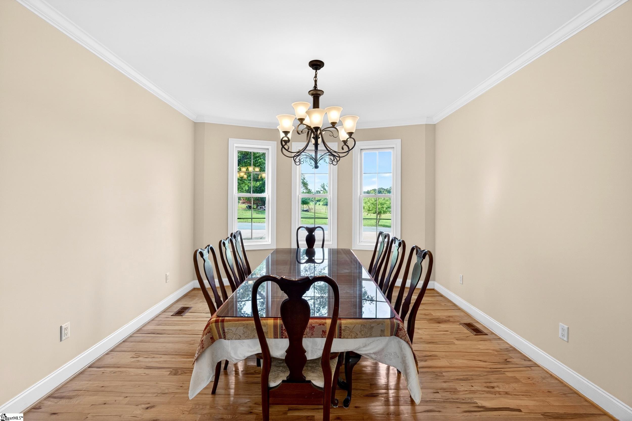 Dining room with a long table, hardwood floors, chandelier, and three windows overlooking a lawn.