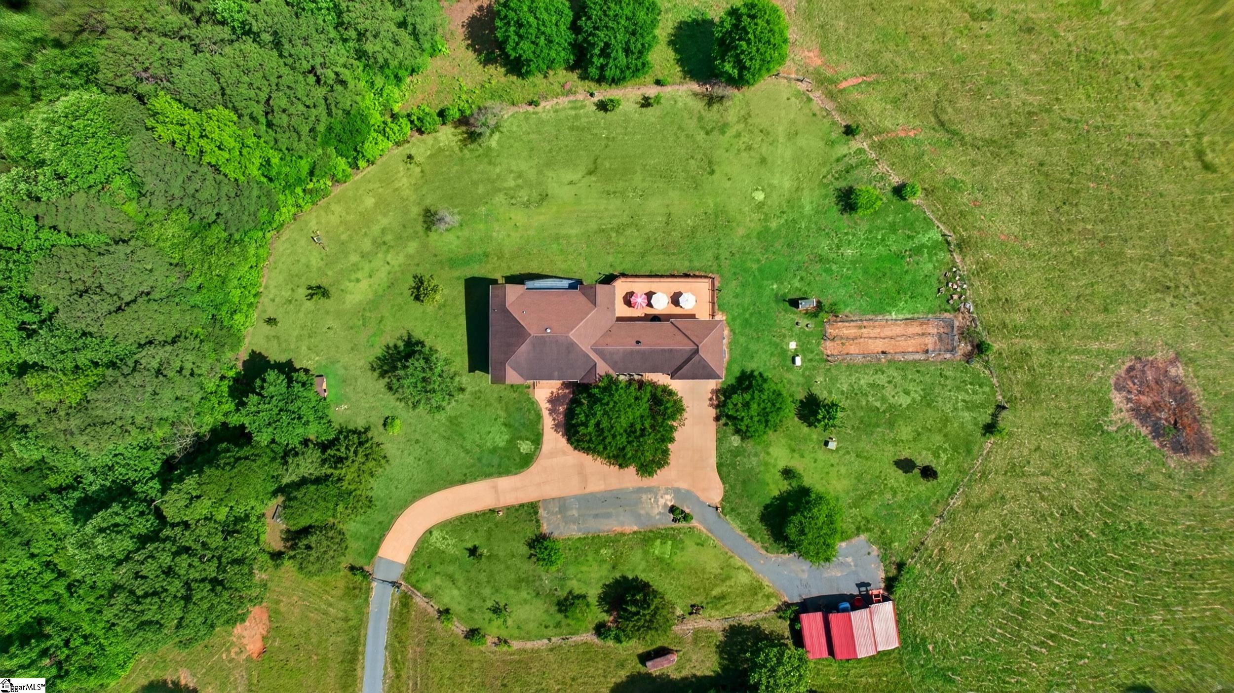 Aerial view of a home surrounded by a broad lawn and trees, with a curved driveway and red-roofed outbuilding.