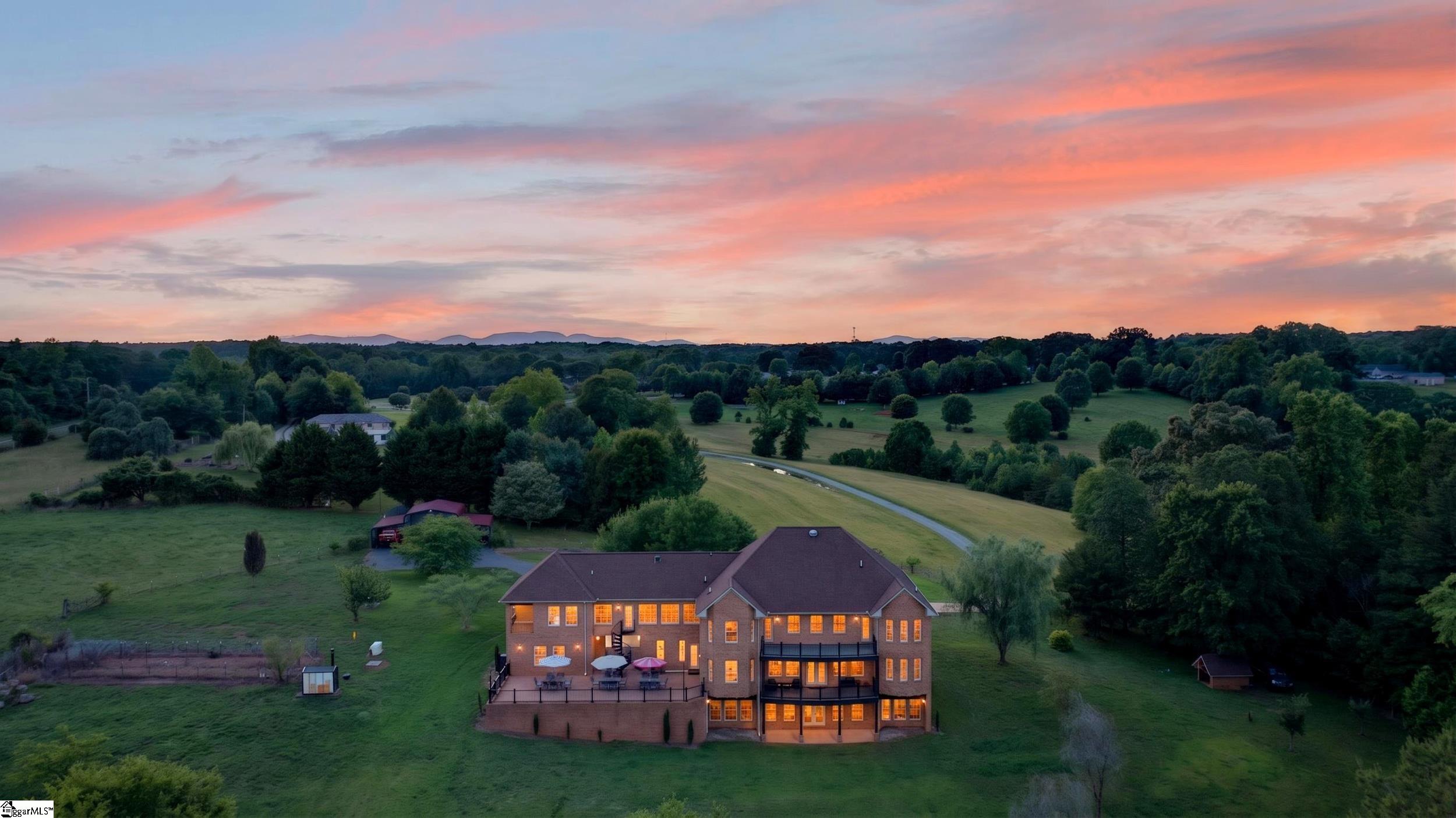 Aerial view of a large brick home with glowing windows and expansive green grounds beneath a colorful dusk sky.