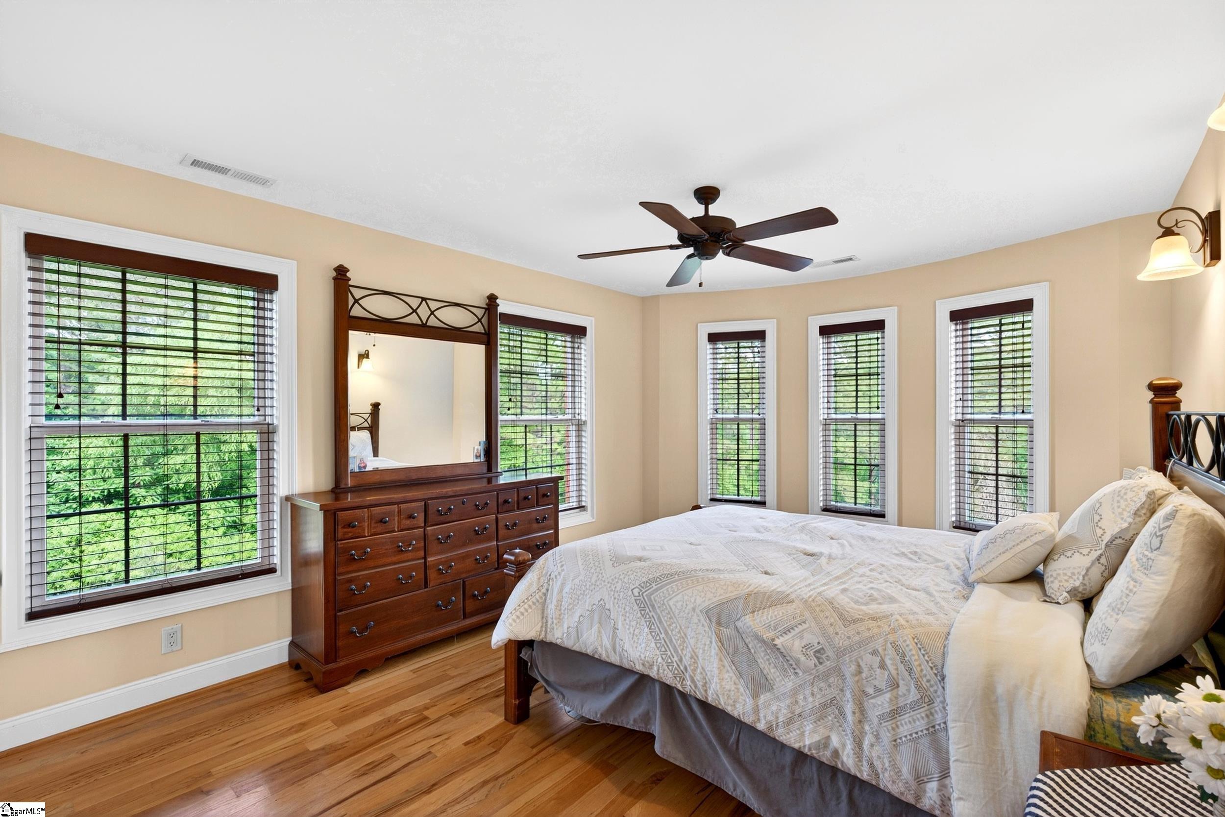 Bedroom with a large bed, dark wood dresser, ceiling fan, and multiple windows overlooking greenery.