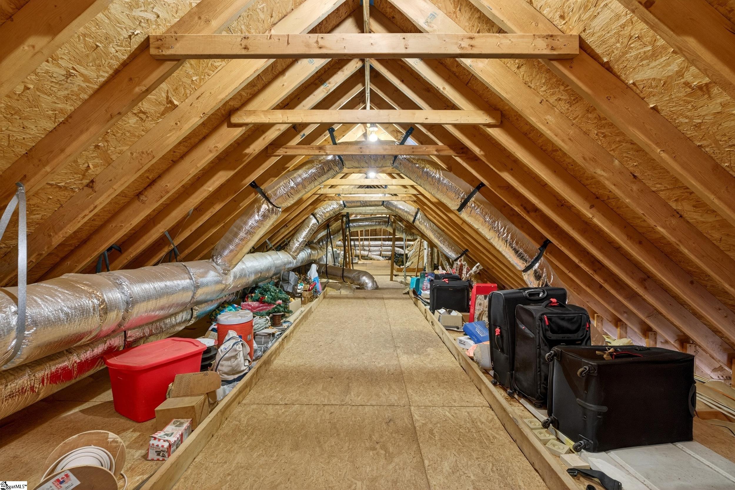 Attic storage area with exposed wooden trusses, silver HVAC ducts, and suitcases lining a plywood walkway.