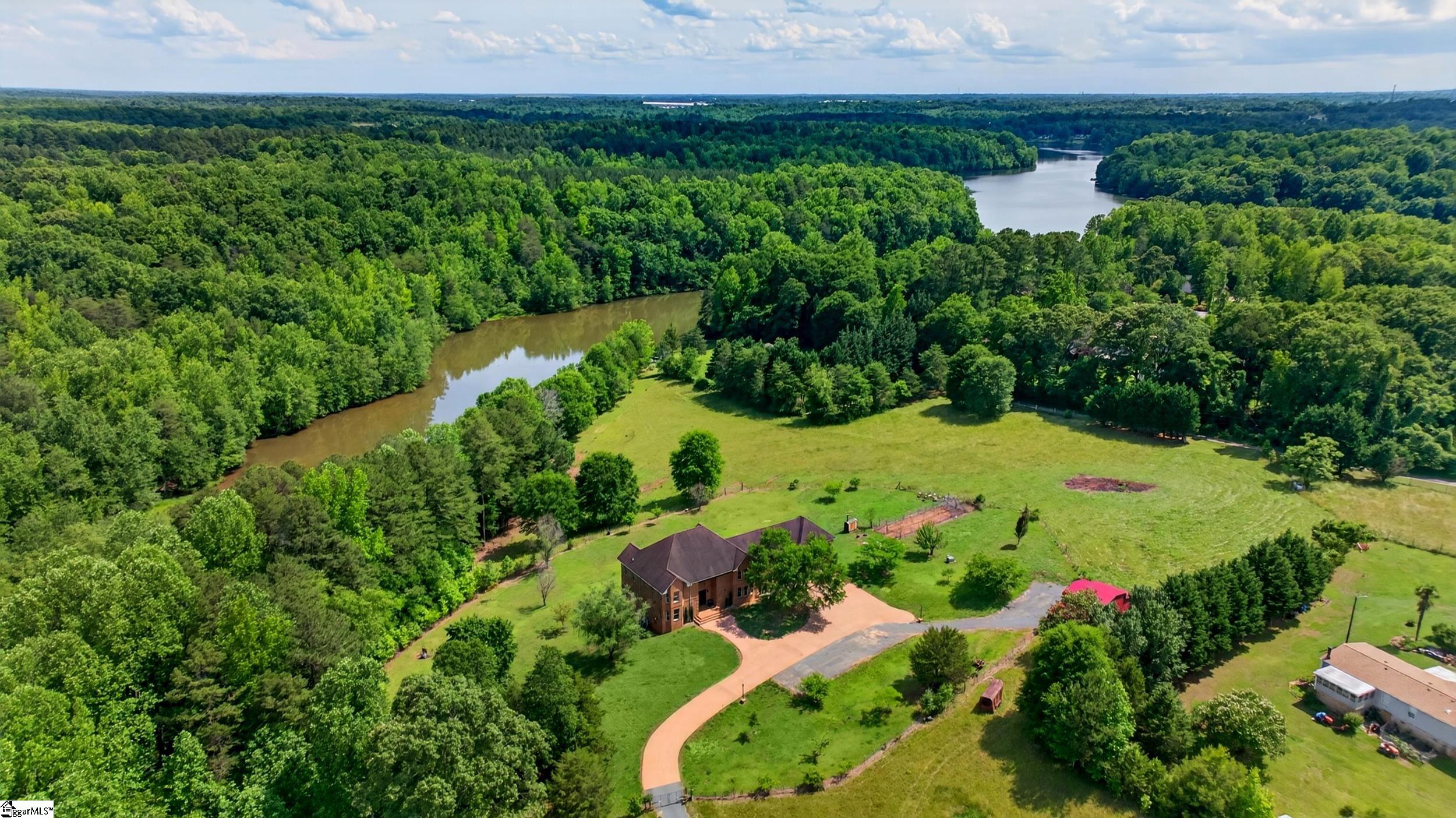 Aerial view of a brick home beside a wooded river, with expansive green lawn and a curving driveway.