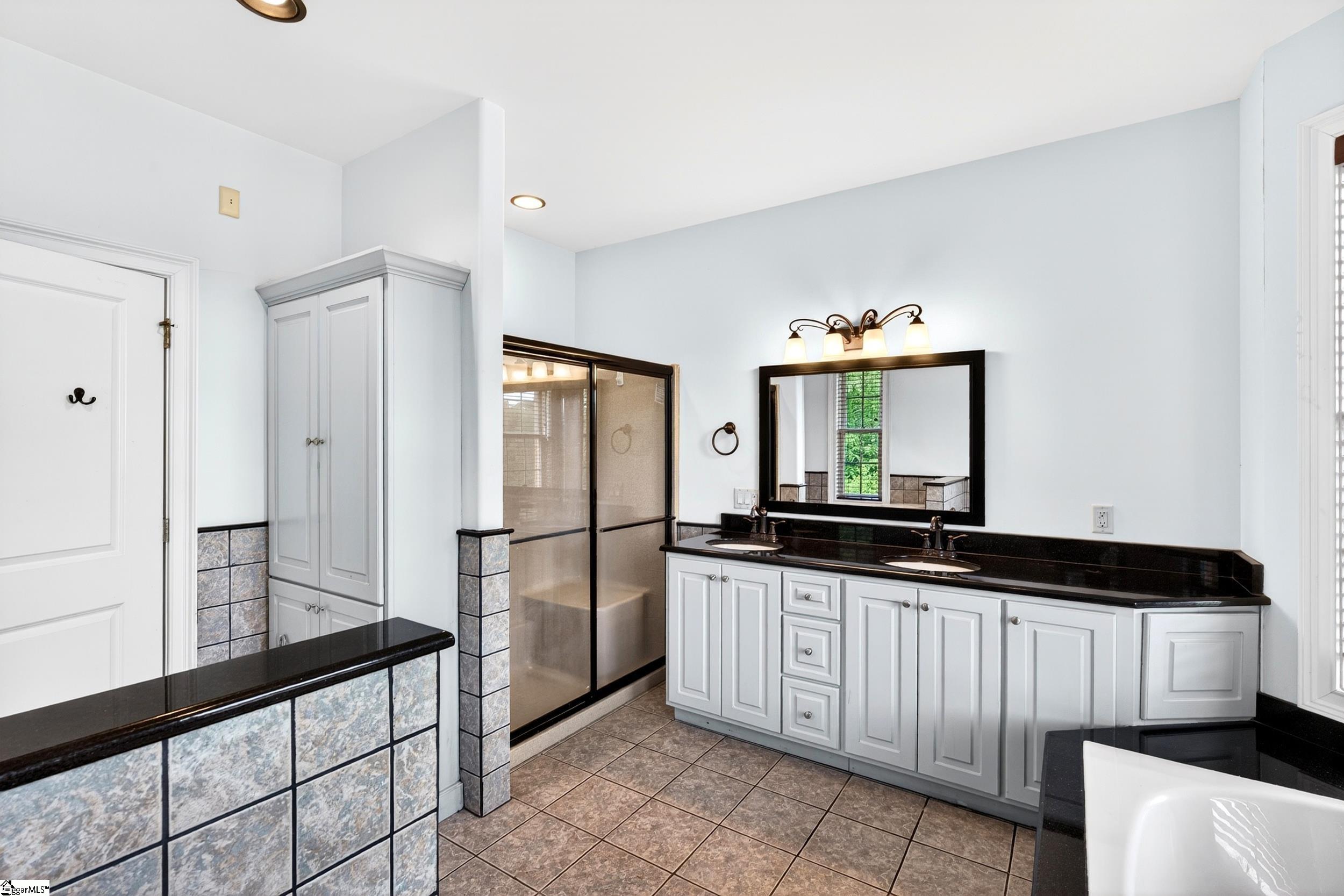 Bathroom with a double-sink vanity, black countertops, glass-enclosed shower, and partially visible soaking tub.