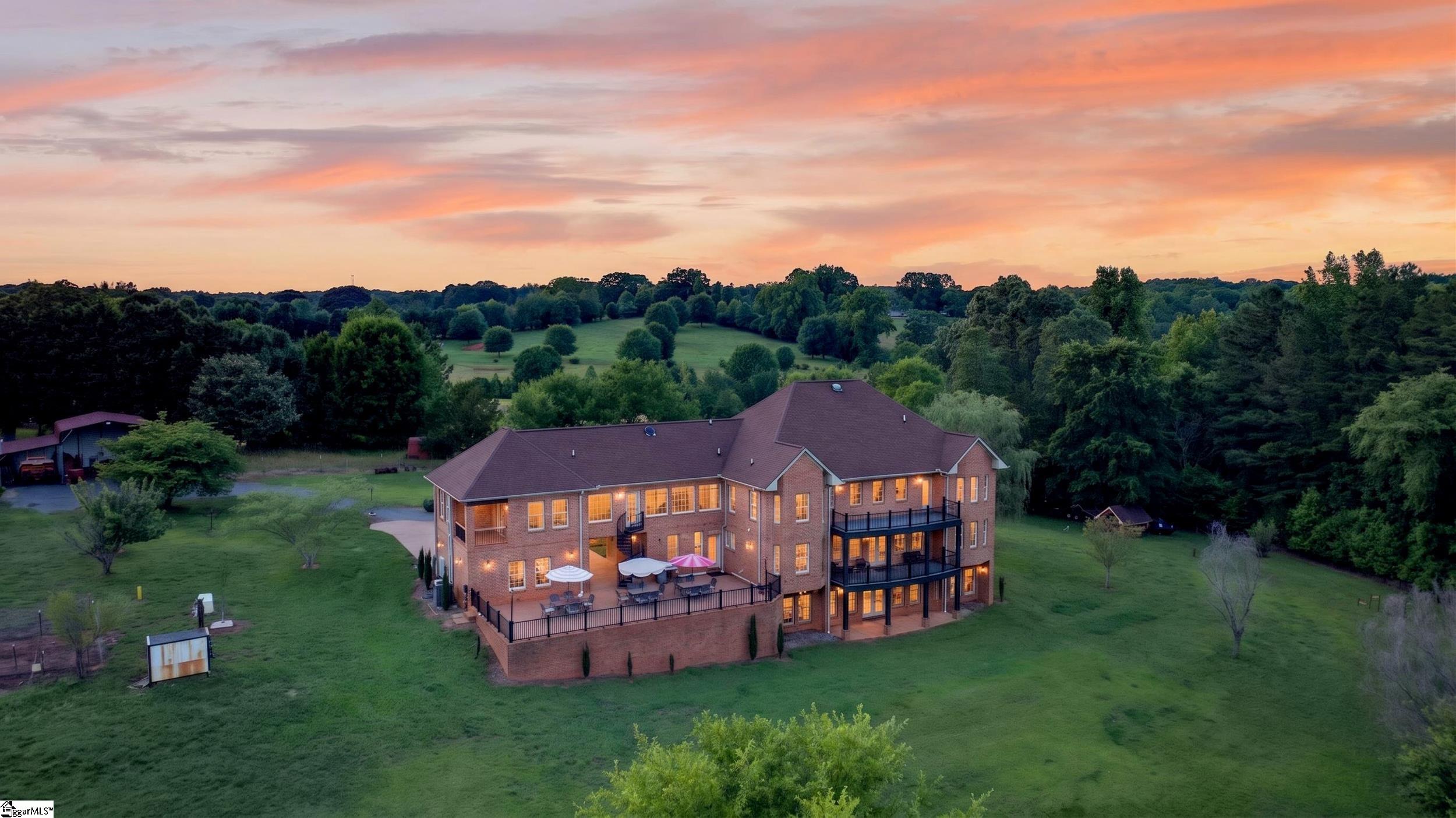 Aerial view of a large brick home with a broad lawn and an illuminated patio at dusk.